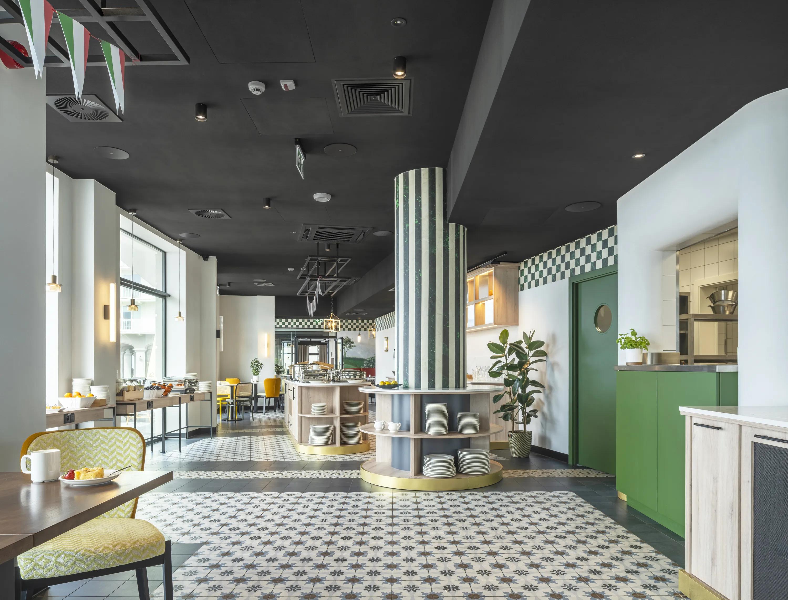Bright, modern café interior with patterned floor tiles, black suspended ceiling, green and yellow chairs, wall art, hanging lights, and plants for decoration.