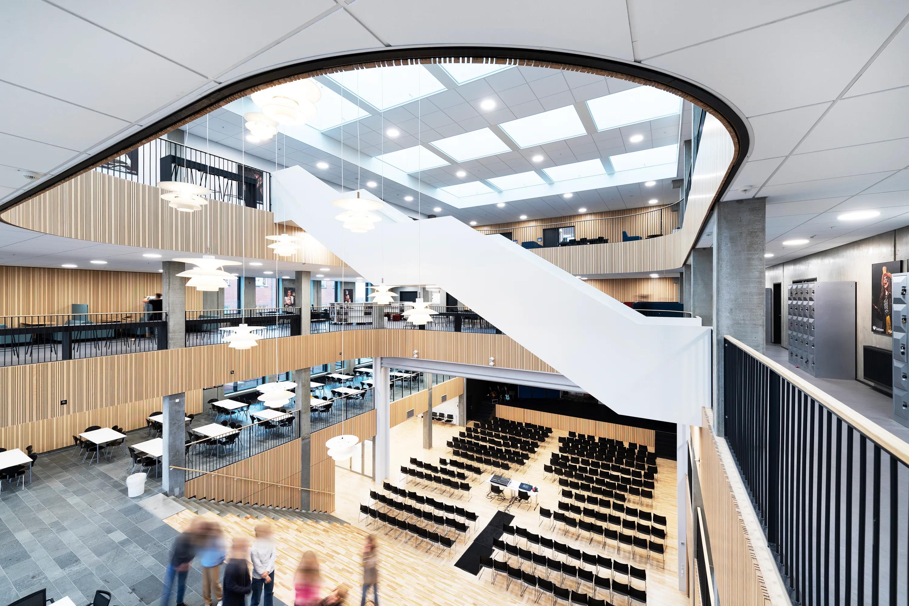 A spacious, multi-storey school atrium featuring white acoustic ceiling tiles and large skylights above open walkways, tiered seating, and a central hall arranged for an assembly