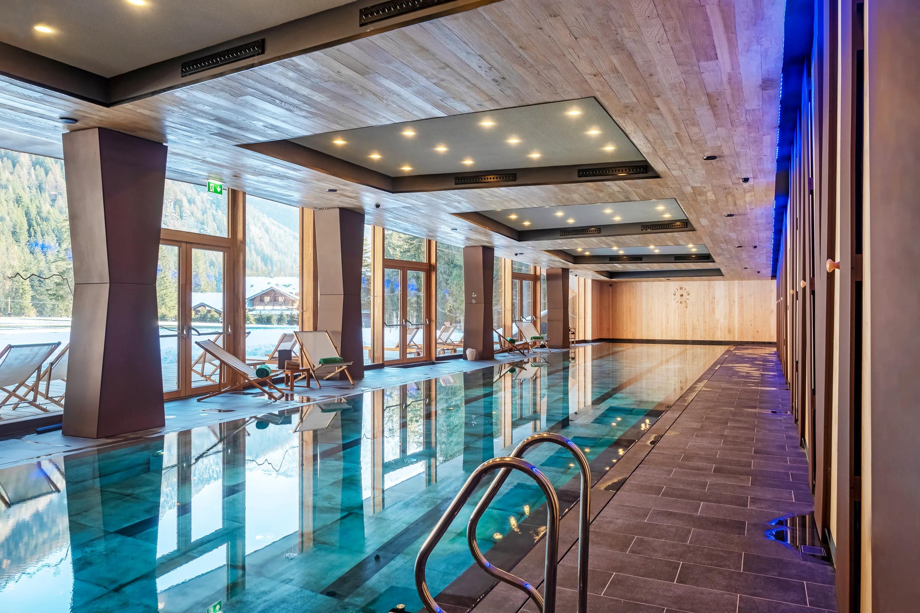 Indoor swimming pool with large floor-to-ceiling windows, wooden ceiling panels with integrated lighting, and lounge chairs overlooking a snowy mountain landscape.
