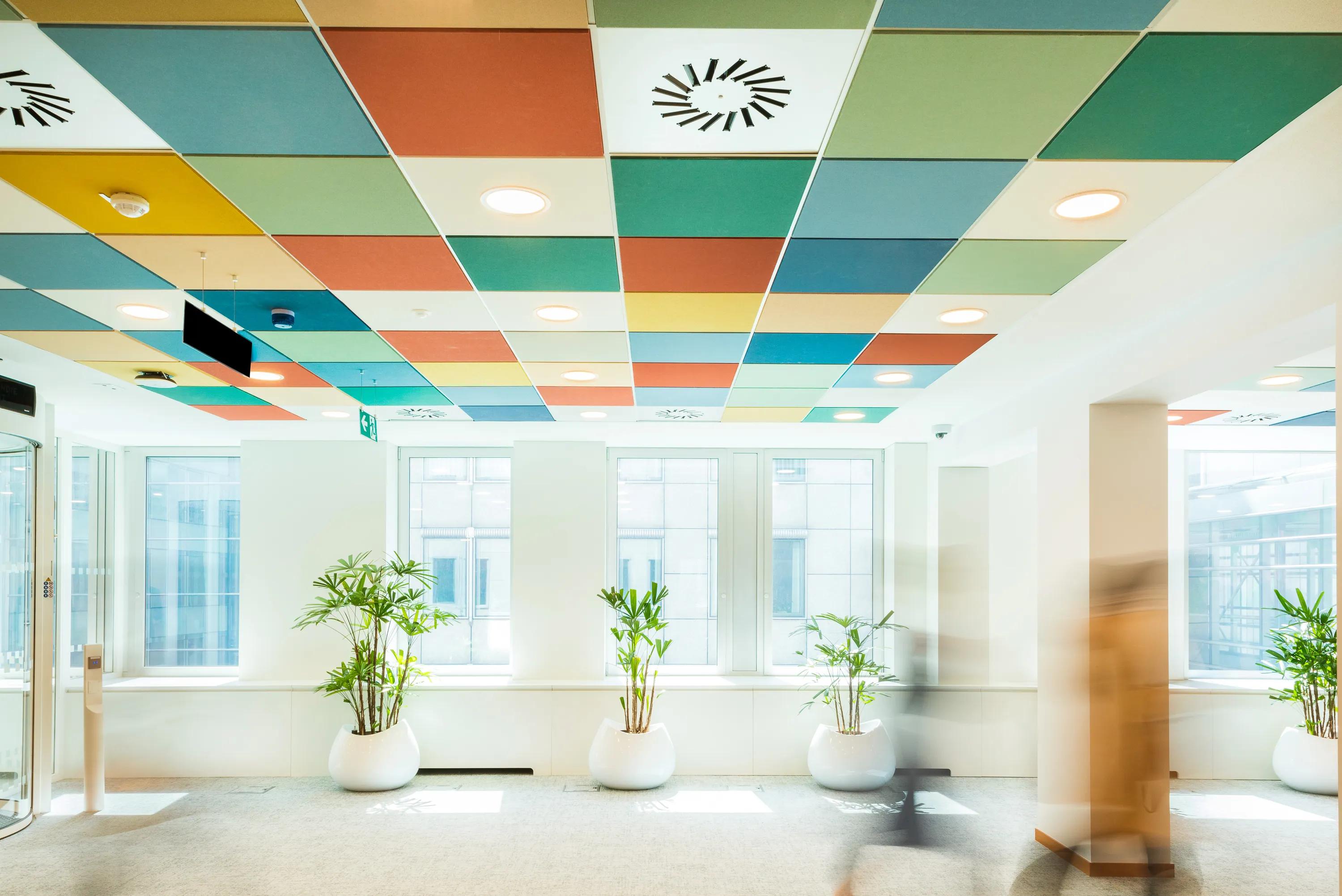 Bright lobby with colorful tiled ceiling, large windows, and white planters with tall green plants. Blurred figures walk through the space.