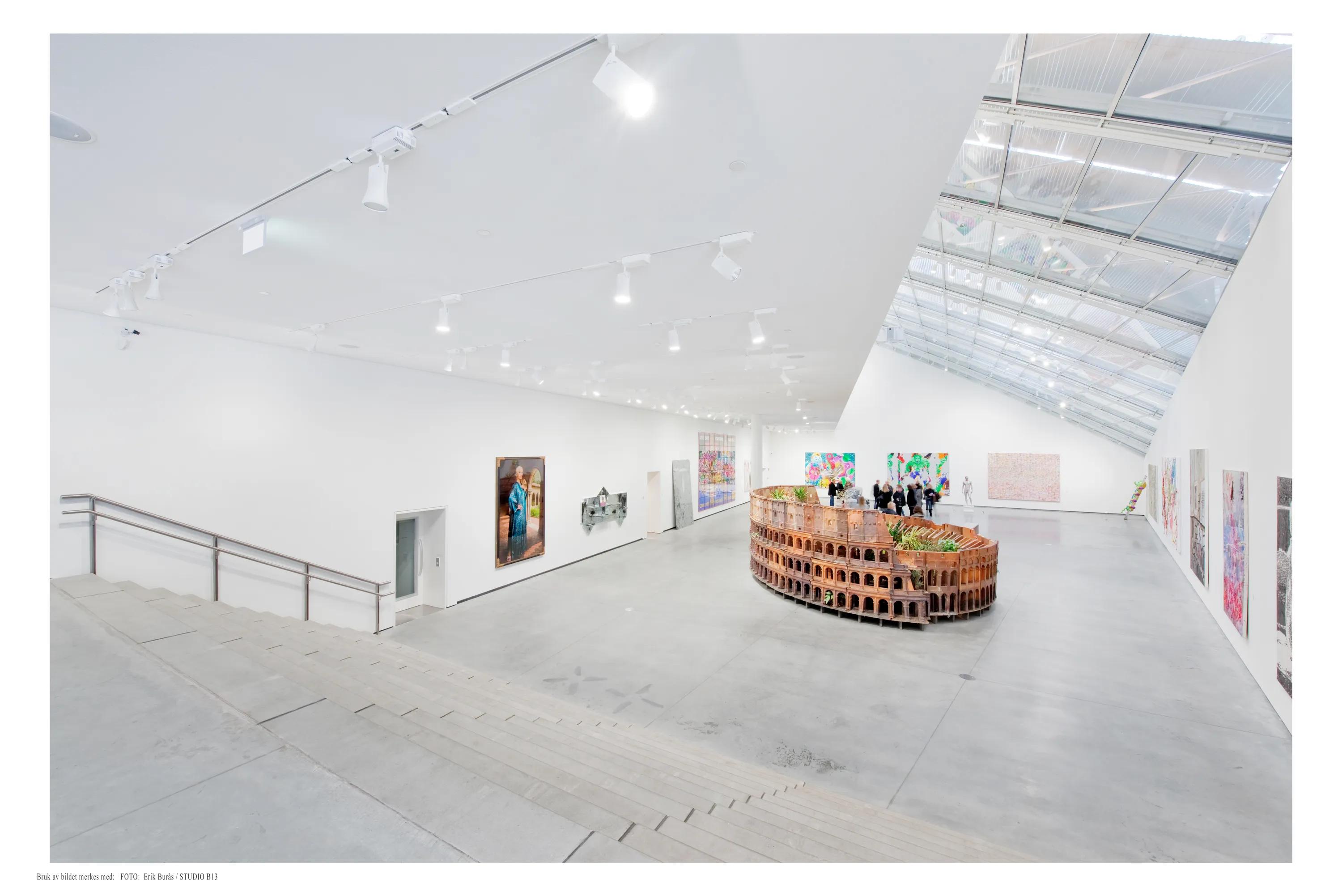 Large contemporary art gallery with a white acoustic ceiling, skylight roof section, track lighting, and colourful artworks displayed around a central sculptural installation.