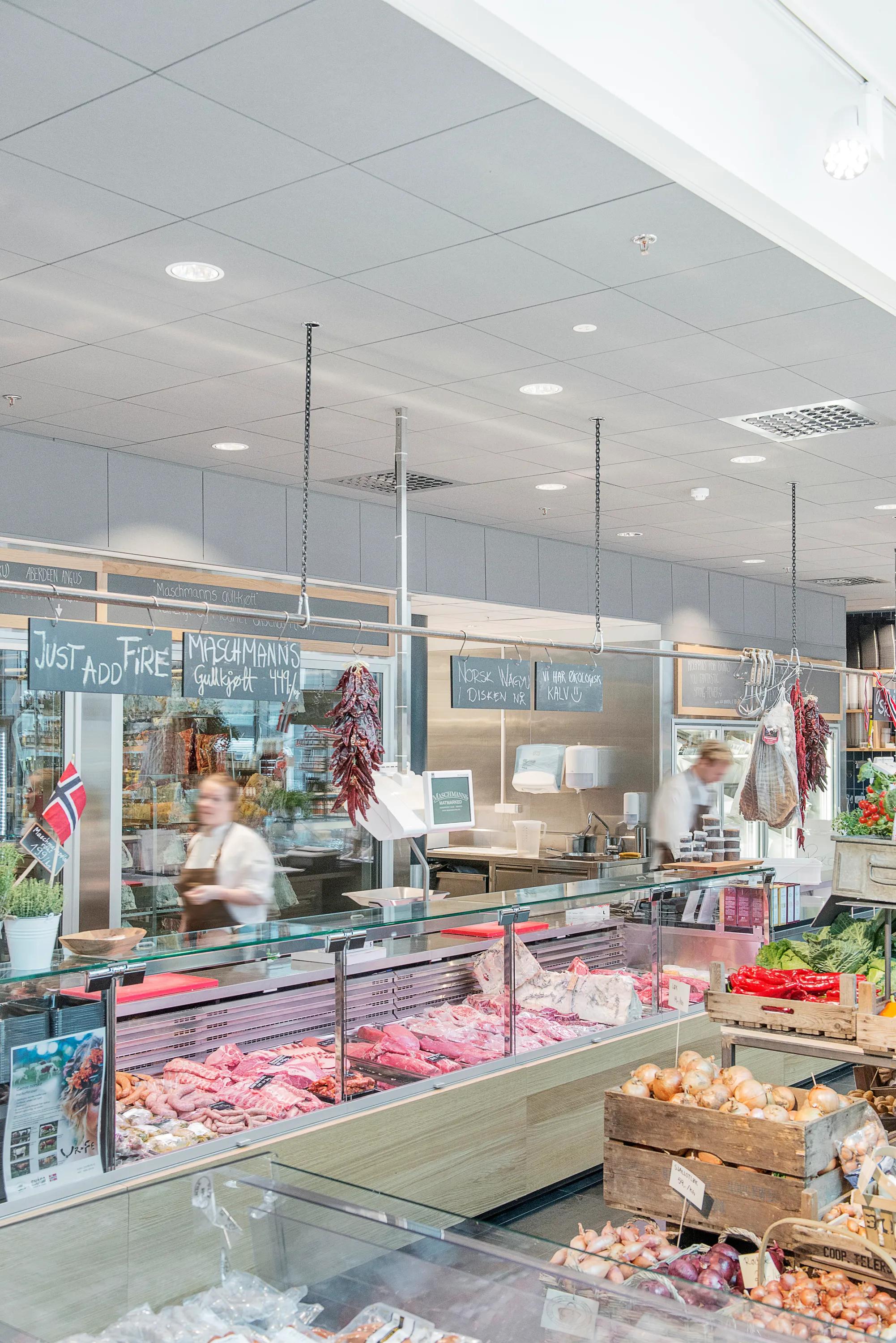 Butcher counter at a food market with refrigerated meat displays and a grey acoustic tile ceiling above the service area.