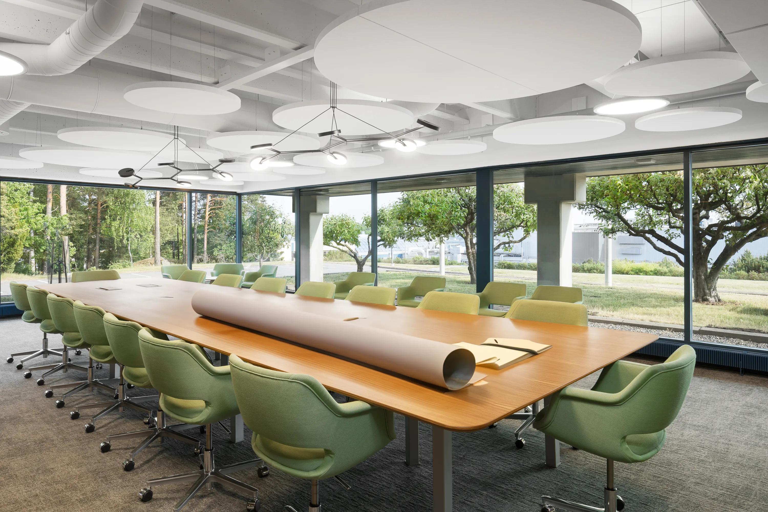 Modern conference room with a long wooden table, green swivel chairs, large windows, and circular ceiling lights and white floating office ceiling, overlooking a garden.