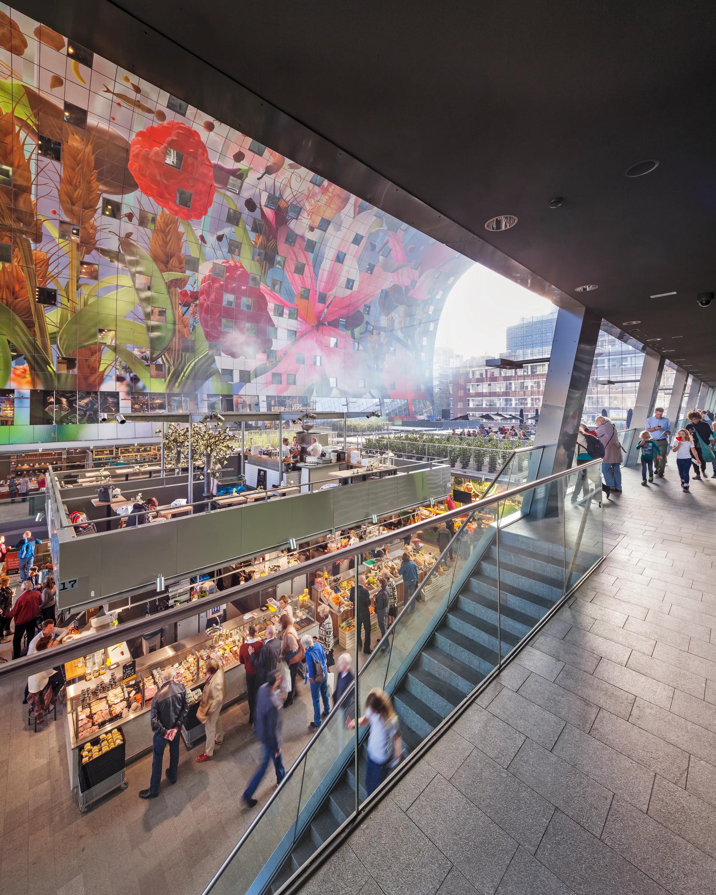 Interior view of Markthal Rotterdam with food market stalls, visitors, and the large colourful ceiling artwork spanning the vaulted structure.