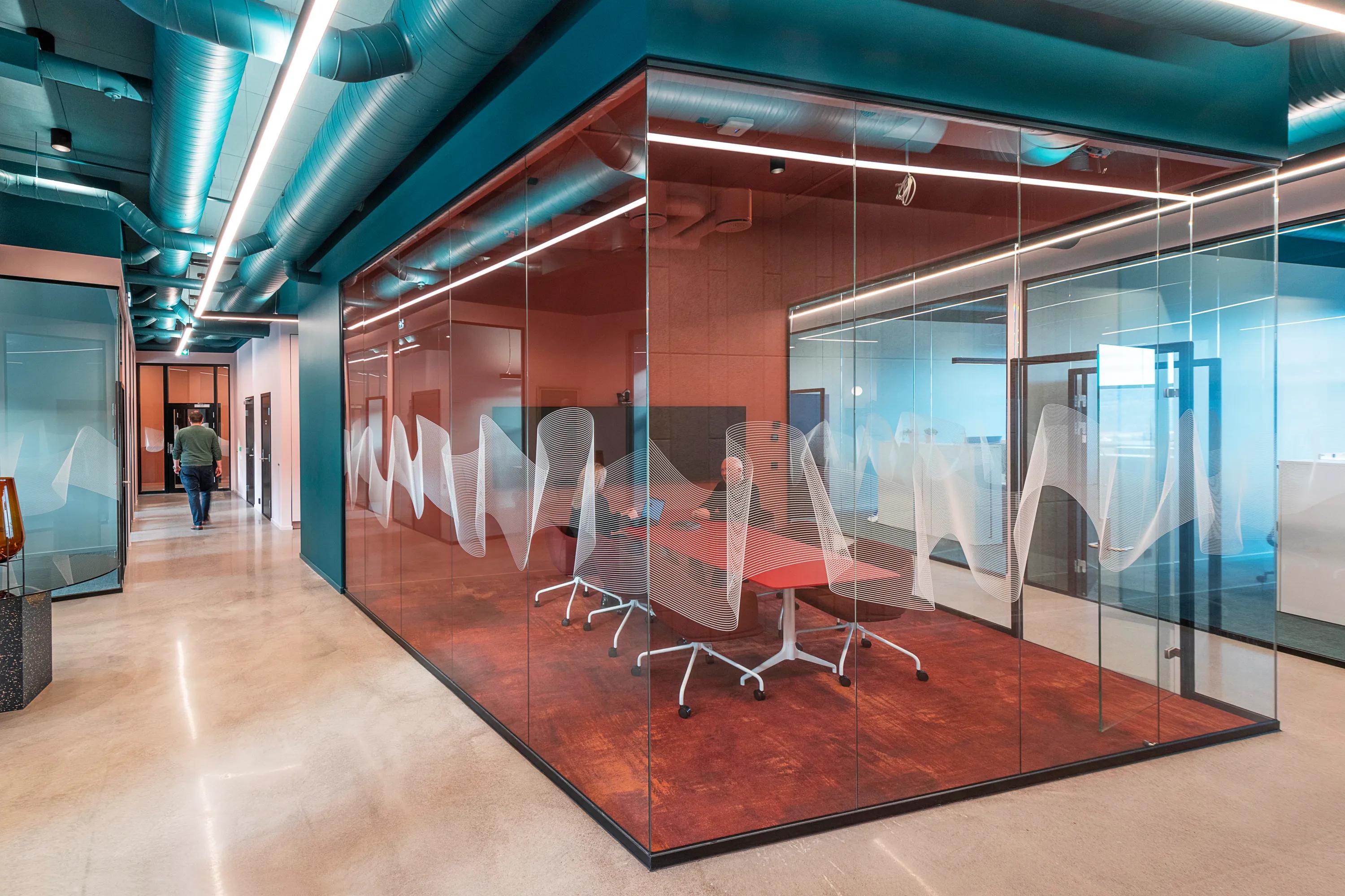 Office corridor with exposed services and acoustic ceiling elements, glass meeting rooms with coloured finishes, and a collaborative workspace visible through transparent partitions.