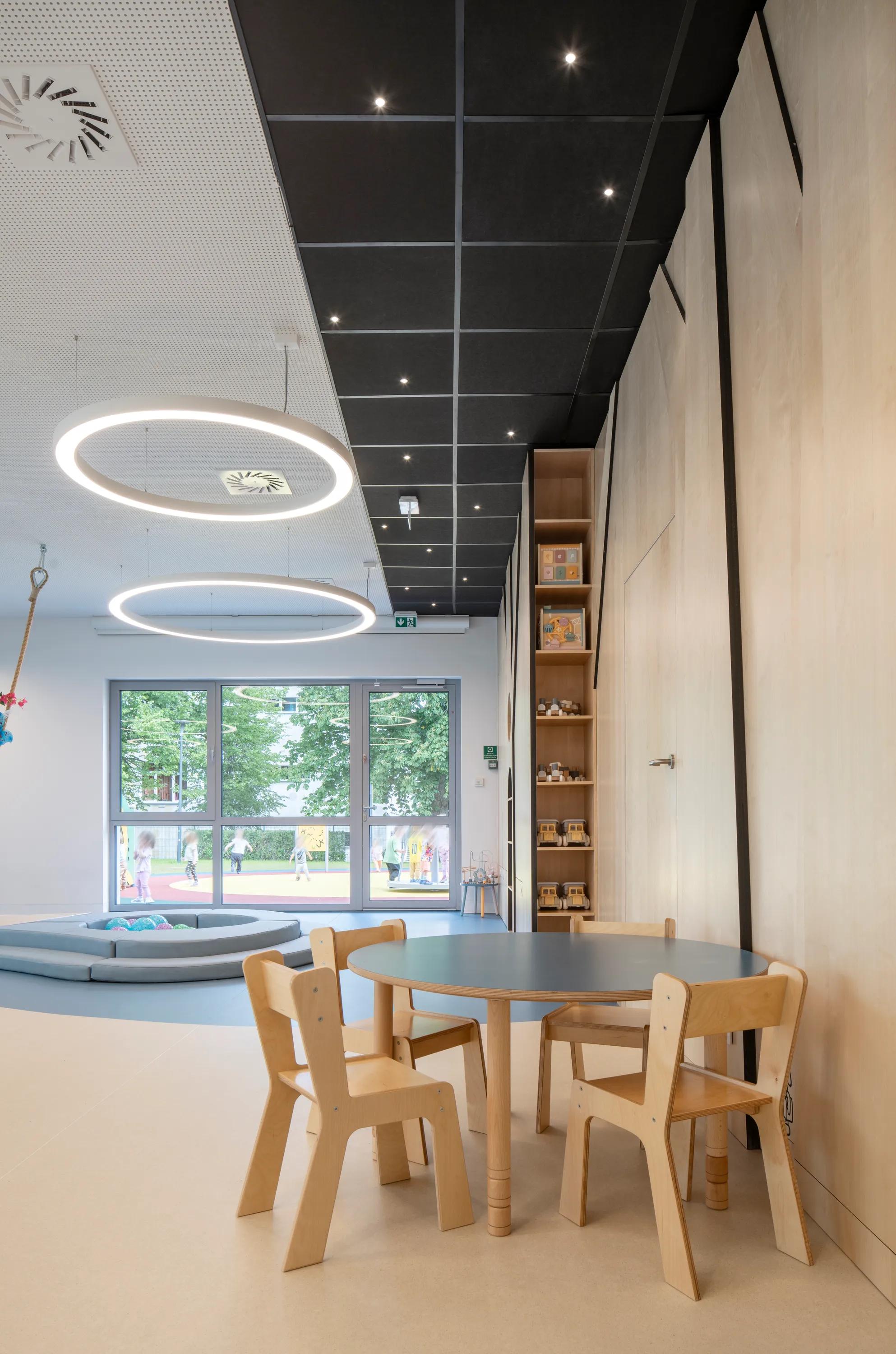 A light-filled playroom featuring black acoustic ceiling tiles with tiny integrated spotlights, large circular pendant lights, and a children’s table set beside tall windows.