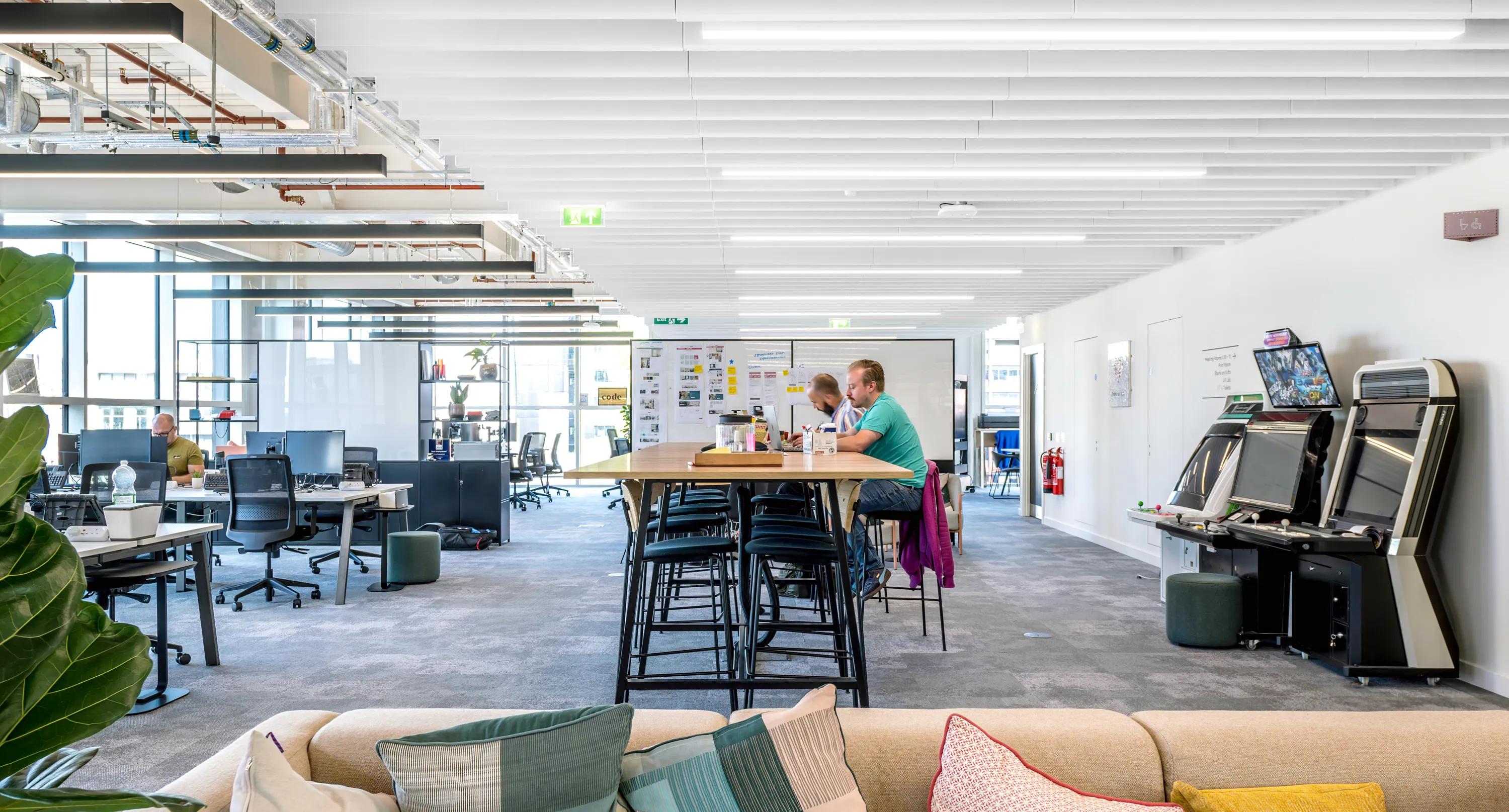 Open-plan office workspace with desks, collaboration tables, and white acoustic baffle ceiling panels above the interior.