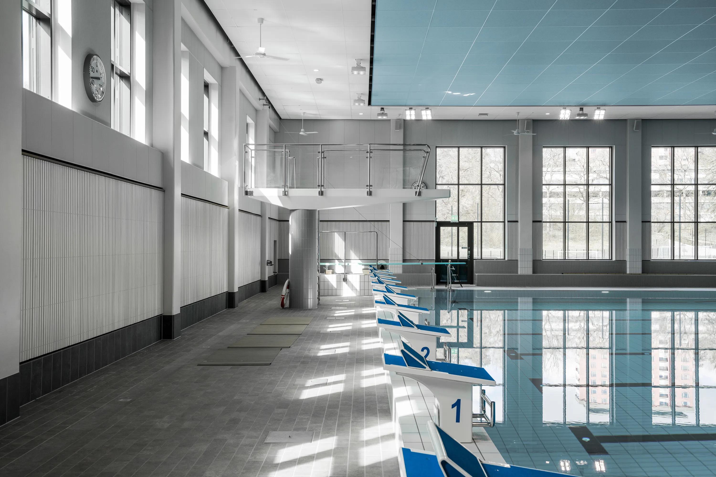 Indoor swimming pool with a blue acoustic ceiling, diving platform, lane markings, and starting blocks aligned along the pool edge in a bright, daylit hall.