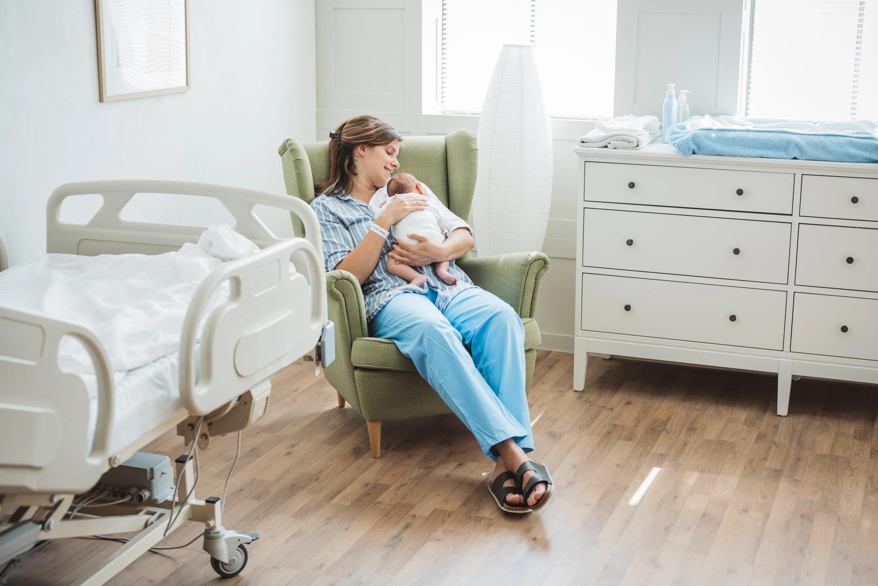 Mother holding newborn in a cozy hospital room with a bed and dresser, sitting in a green armchair by the window.