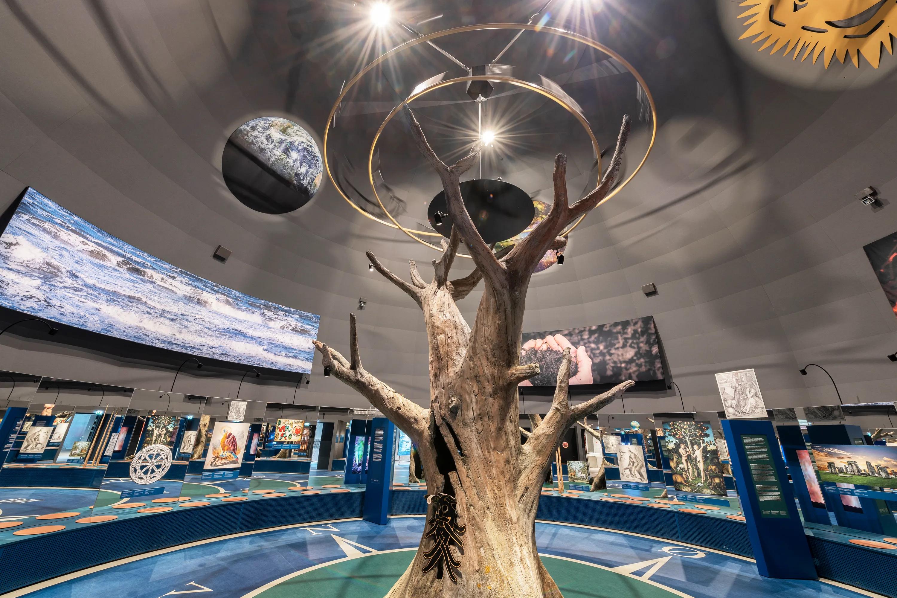Exhibition space at Naturkraft in Ringkøbing with a grey acoustic ceiling, central tree installation, suspended planet models, and interactive displays arranged around the circular gallery.