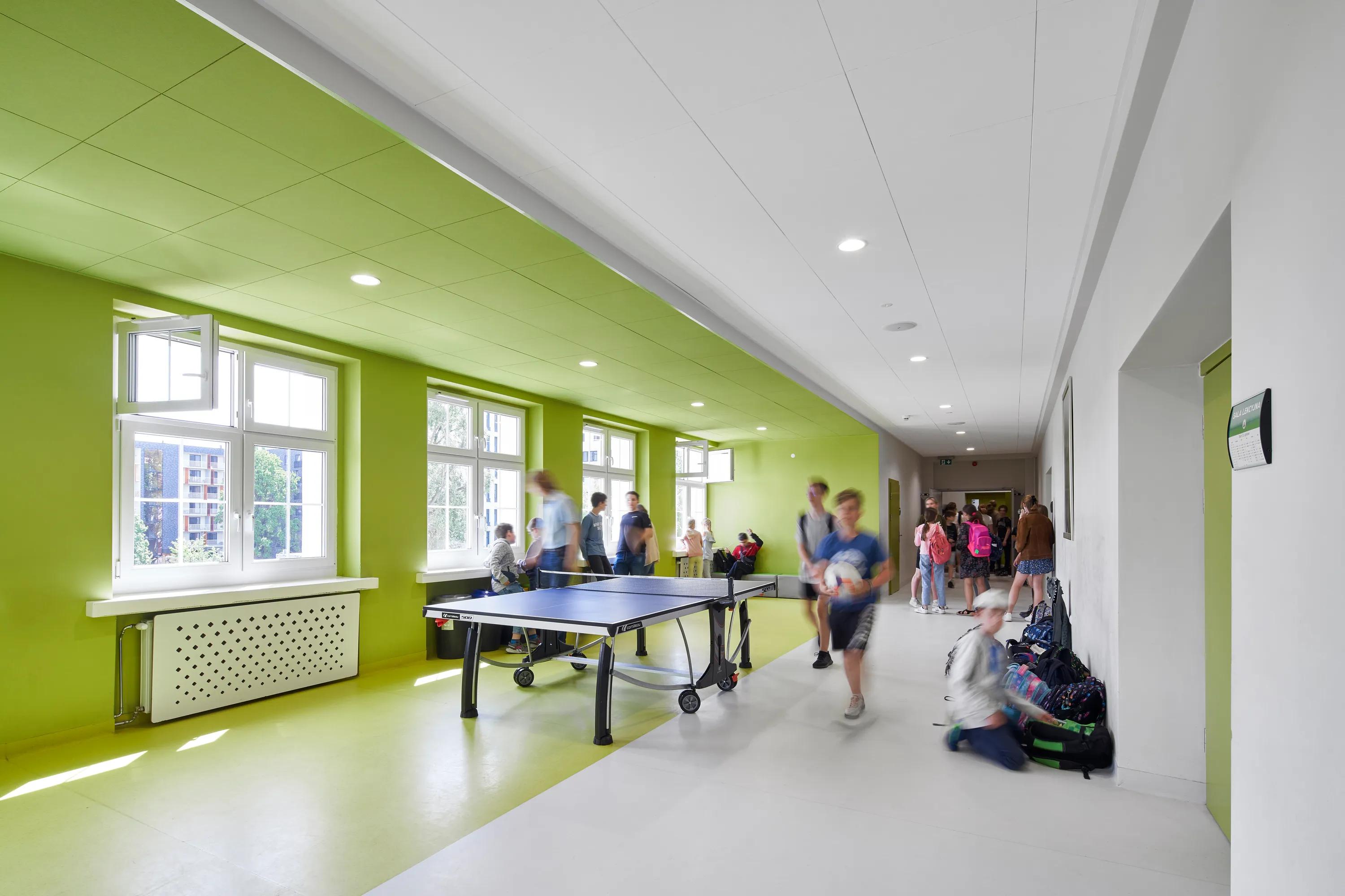 A bright hallway with bright green ceiling and walls, featuring a table tennis setup. Two blurred figures walk by, suggesting motion and activity.