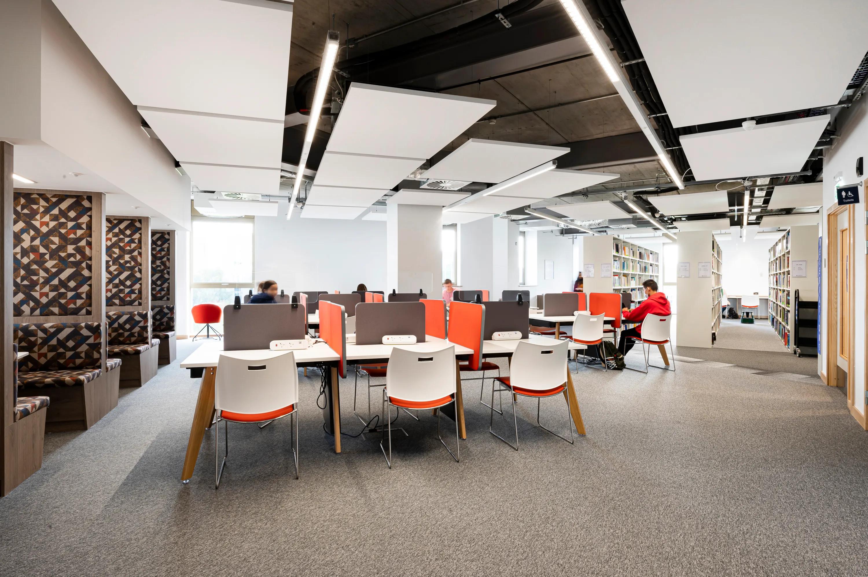 Library study area with suspended white acoustic ceiling panels, desks with study stations, and shelving units arranged in an open, modern learning environment.