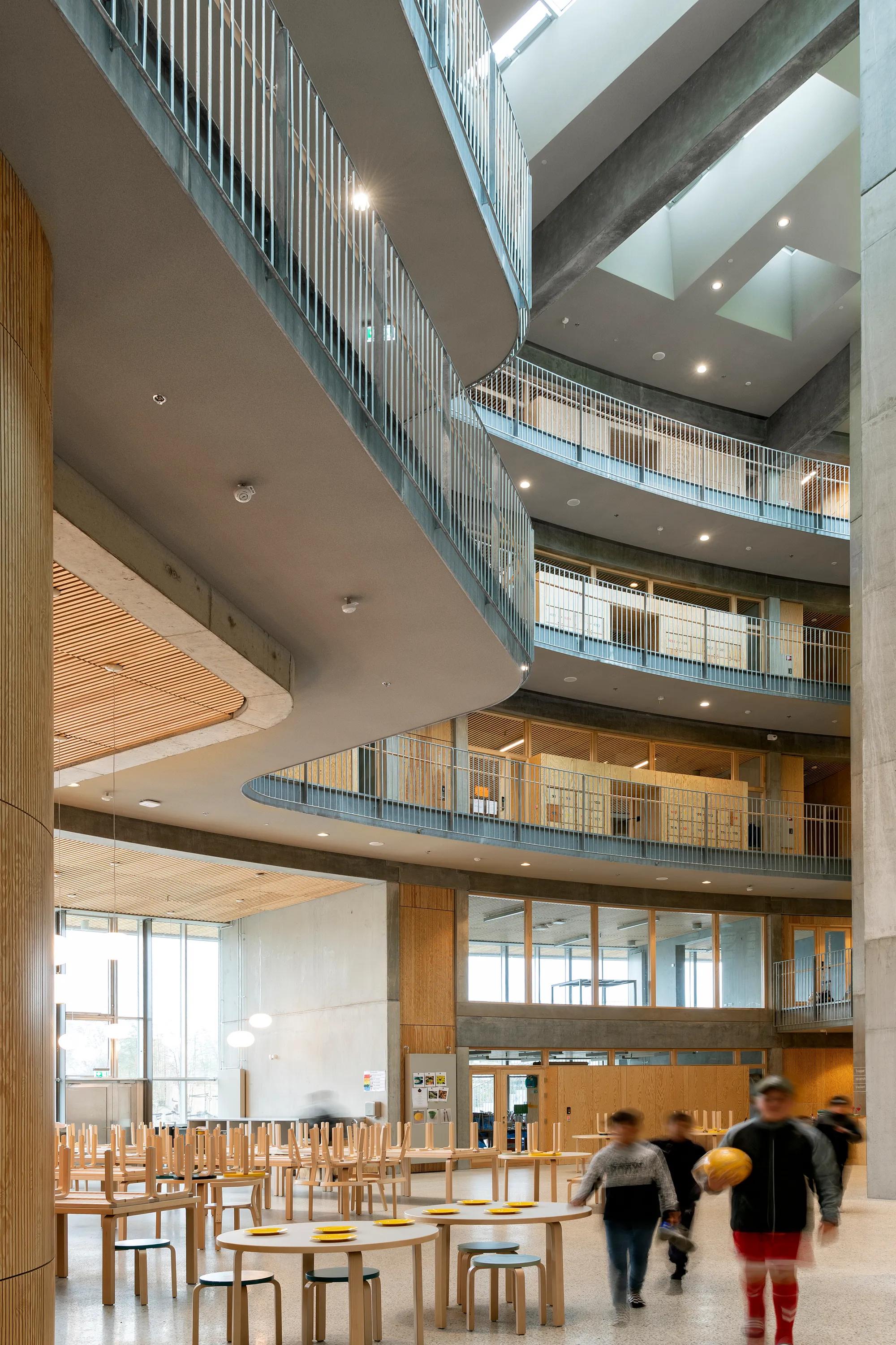 A spacious multi-storey school atrium featuring a seamless monolithic grey acoustic ceiling, curved balcony edges, timber details, and students moving through the bright central space.
