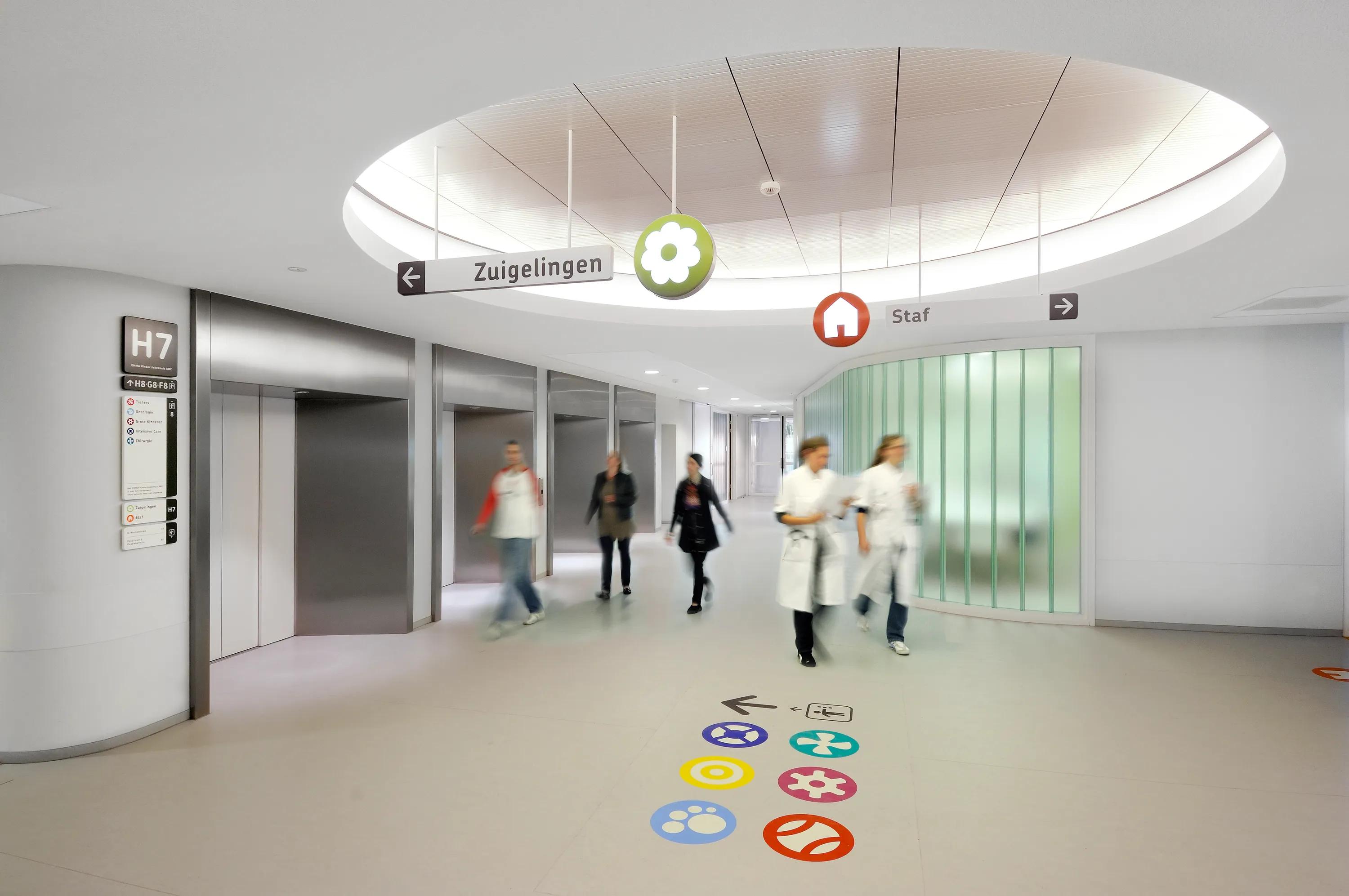Hospital corridor with multiple elevators, seamless monolithic acoustic ceiling feature, wayfinding signage, and medical staff walking through.