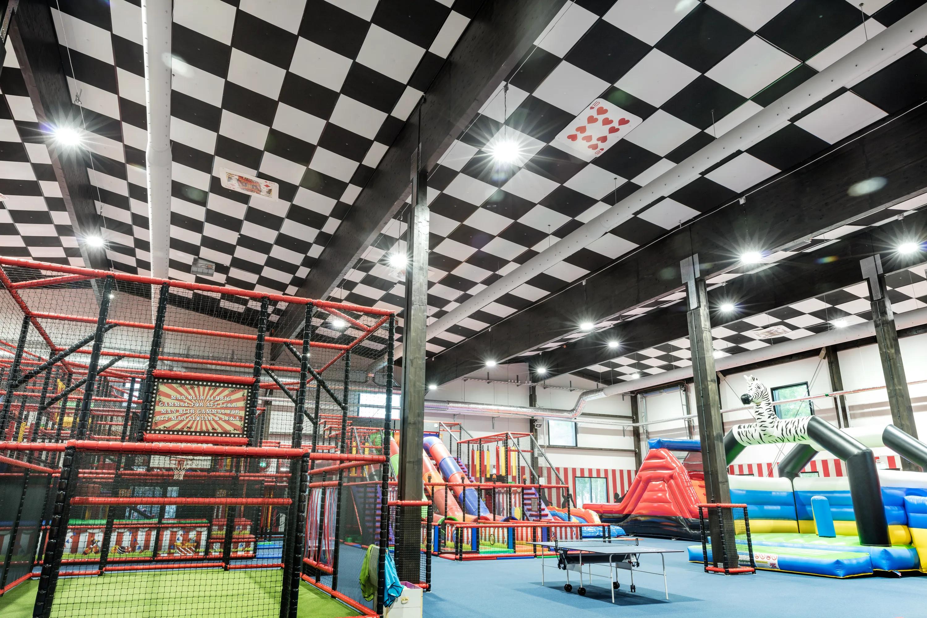 Indoor play centre with a black and white checkerboard acoustic ceiling, climbing frames, slides, trampolines, and inflatable play structures.