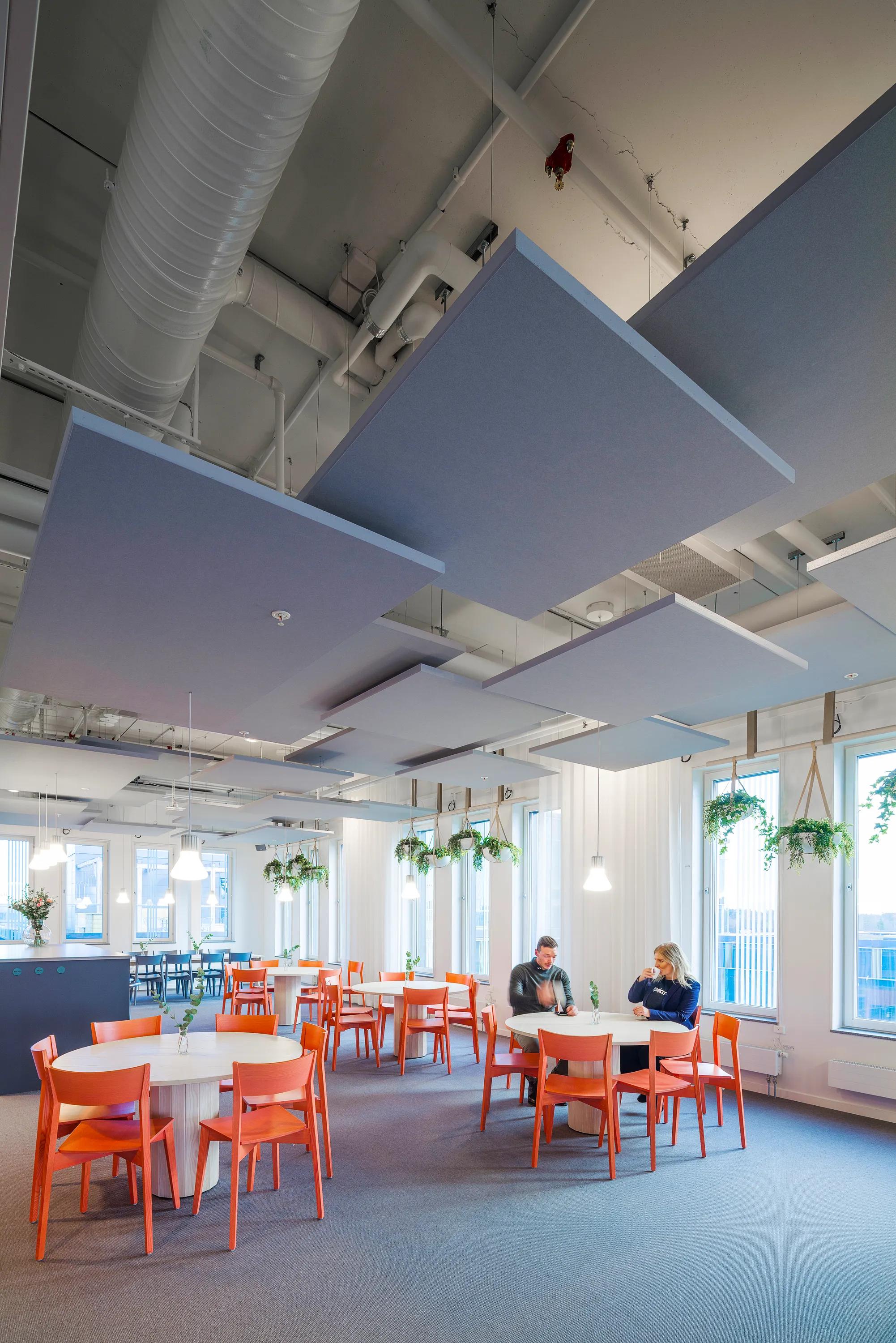Bright cafeteria with orange chairs, a floating raft ceiling, white tables, hanging plants, and acoustic panels. Three people seated at a table in conversation.