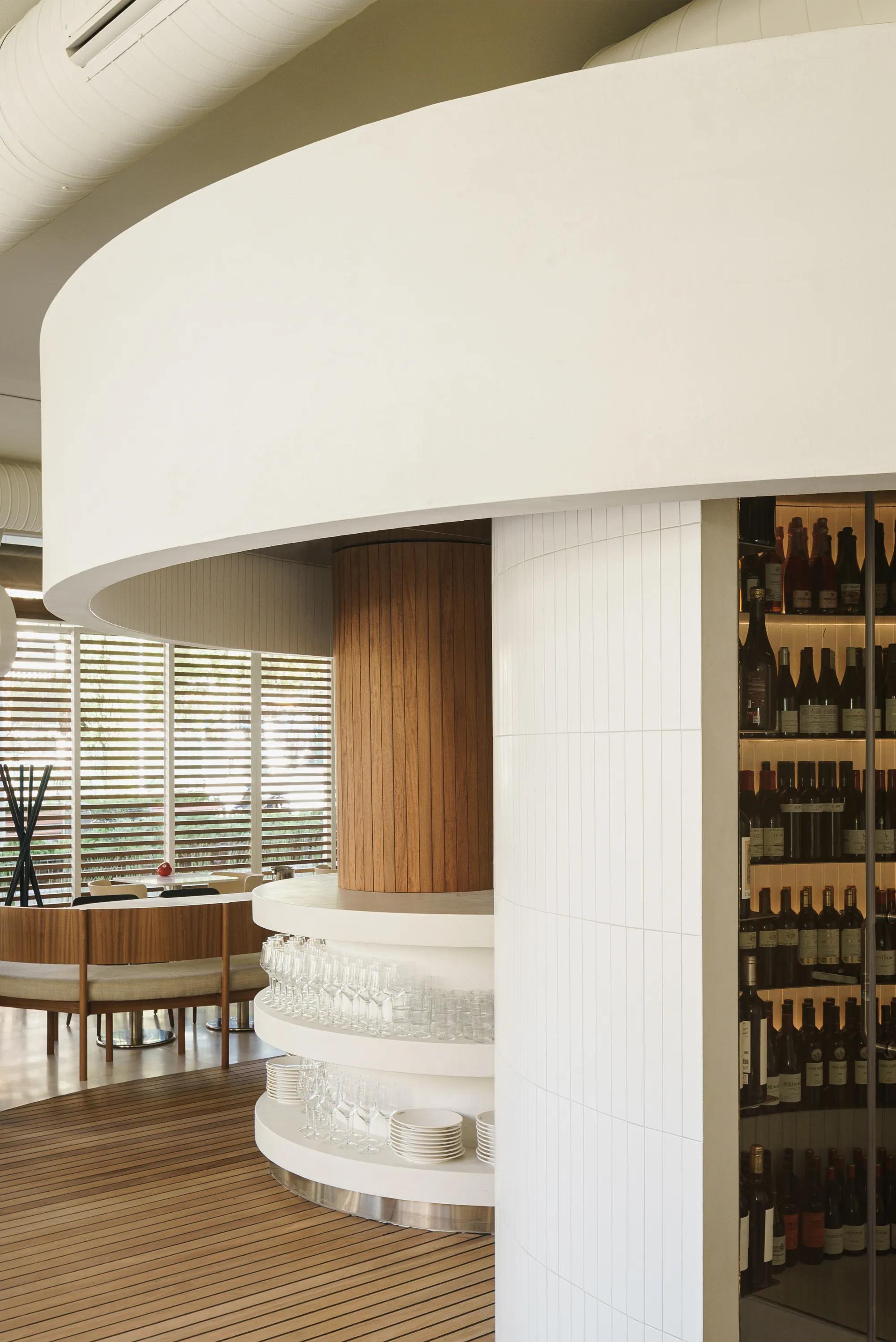 Restaurant interior with a curved monolithic acoustic ceiling above a circular bar, featuring wood columns, wine display shelving, and seating near large windows.