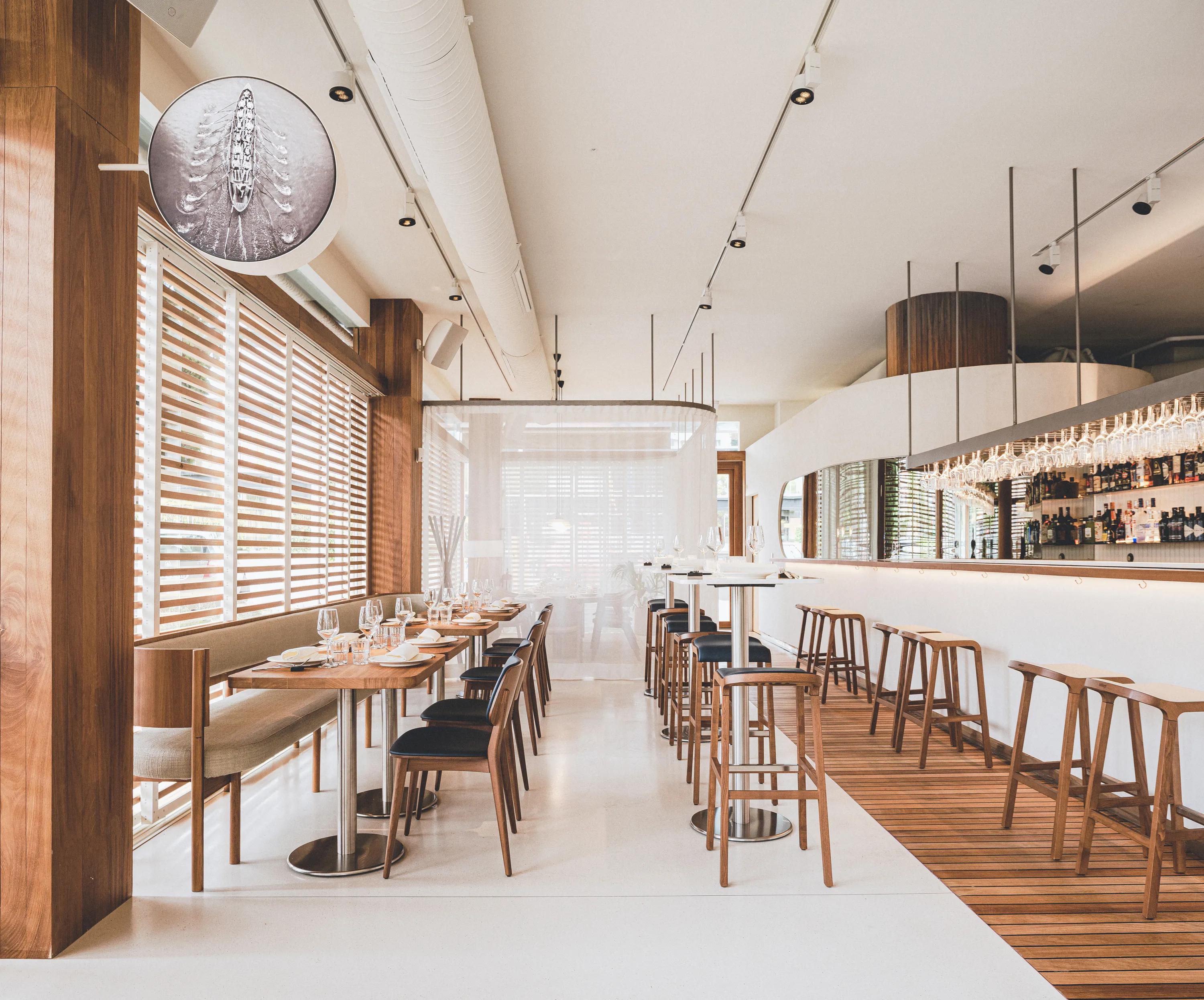 Bright restaurant interior with a white acoustic ceiling, linear track lighting and exposed ventilation duct above wooden dining tables, bar seating and large windows with timber slats.