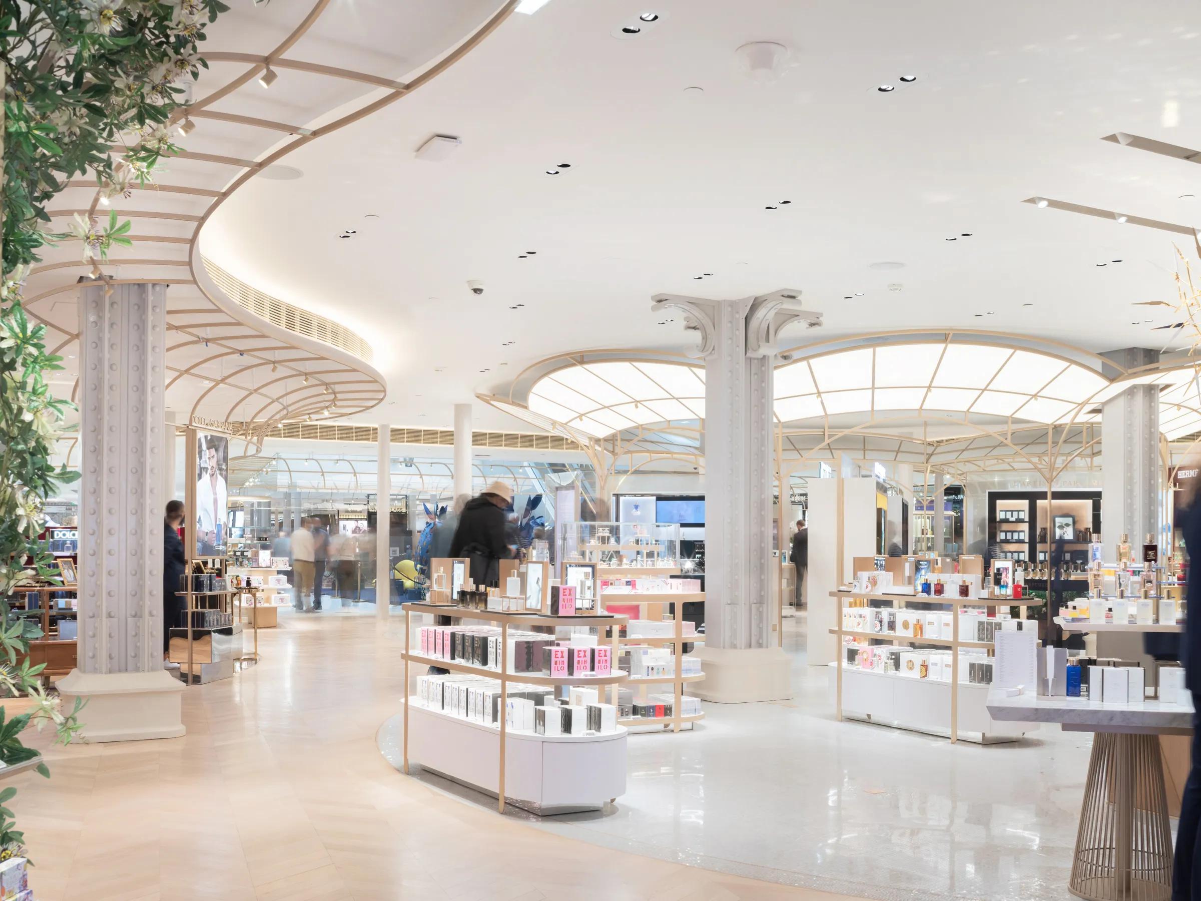 A high-end retail interior featuring a smooth seamless white acoustic ceiling, elegant lattice canopy structures, ornate columns, and island displays filled with perfumes and beauty products.