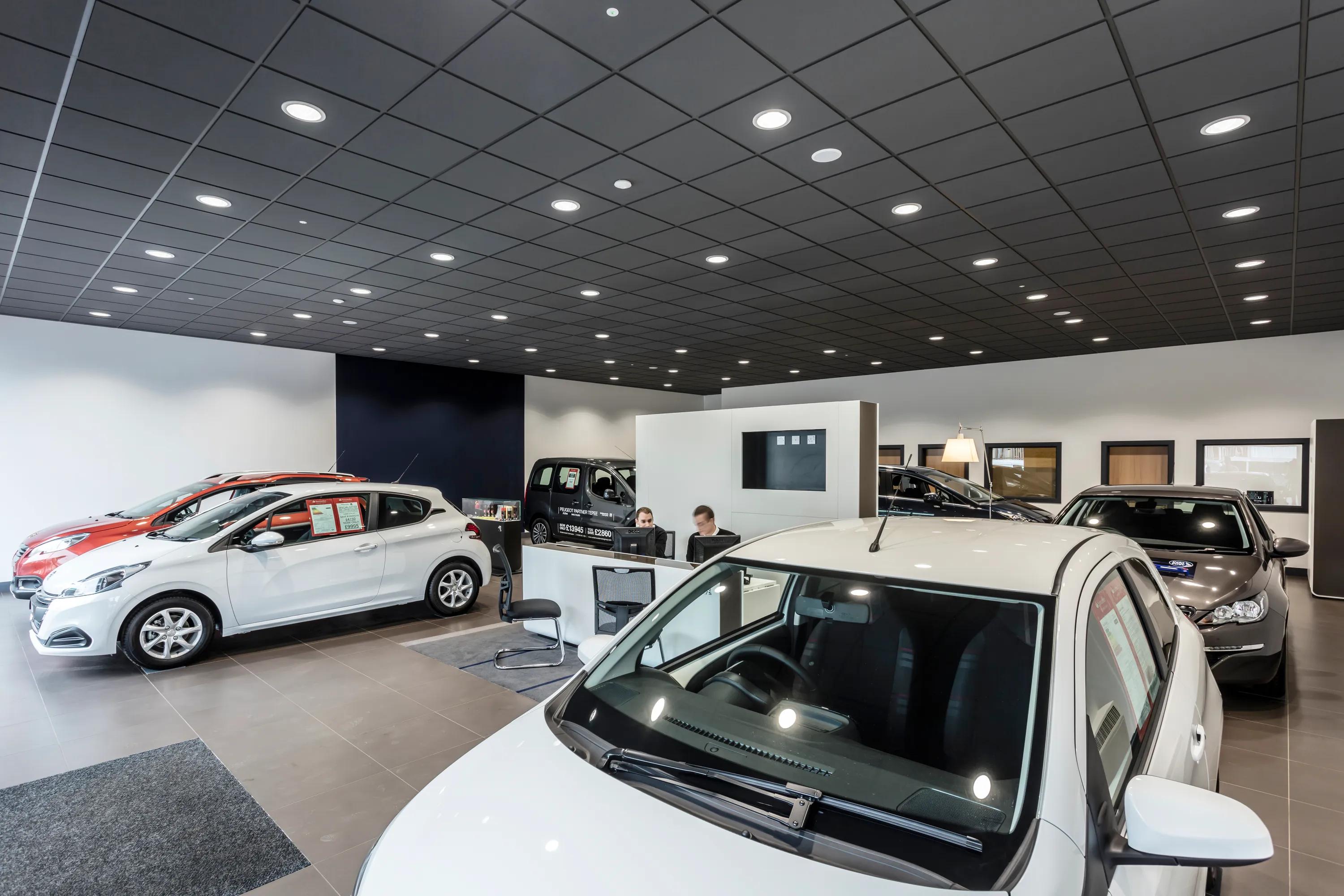 Car dealership showroom with vehicles on display and a black acoustic grid ceiling with recessed lighting above the space.