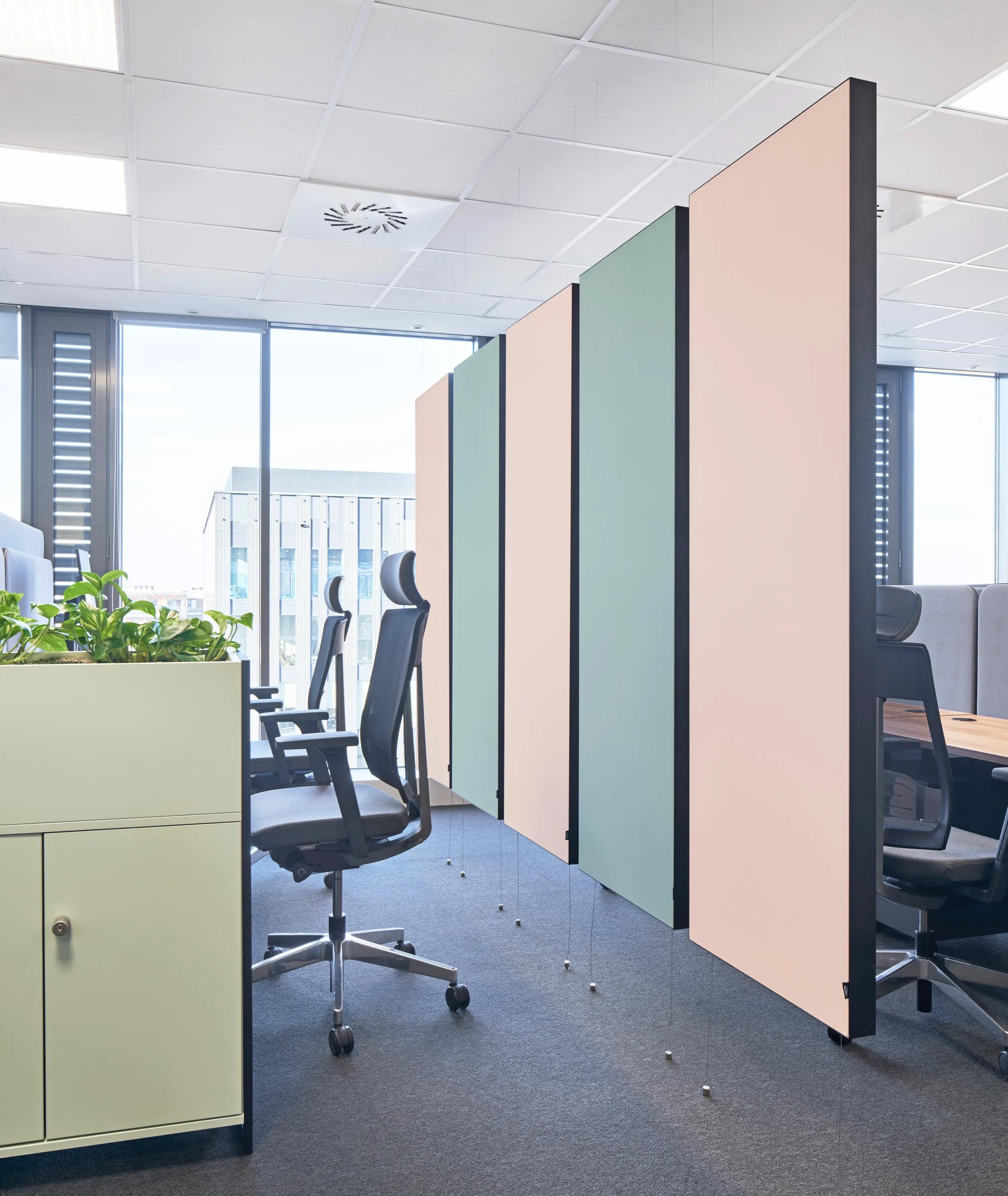Modern office space with suspended pastel acoustic panels, workstation pods, plant-filled modular partitions, and a white grid acoustic ceiling.