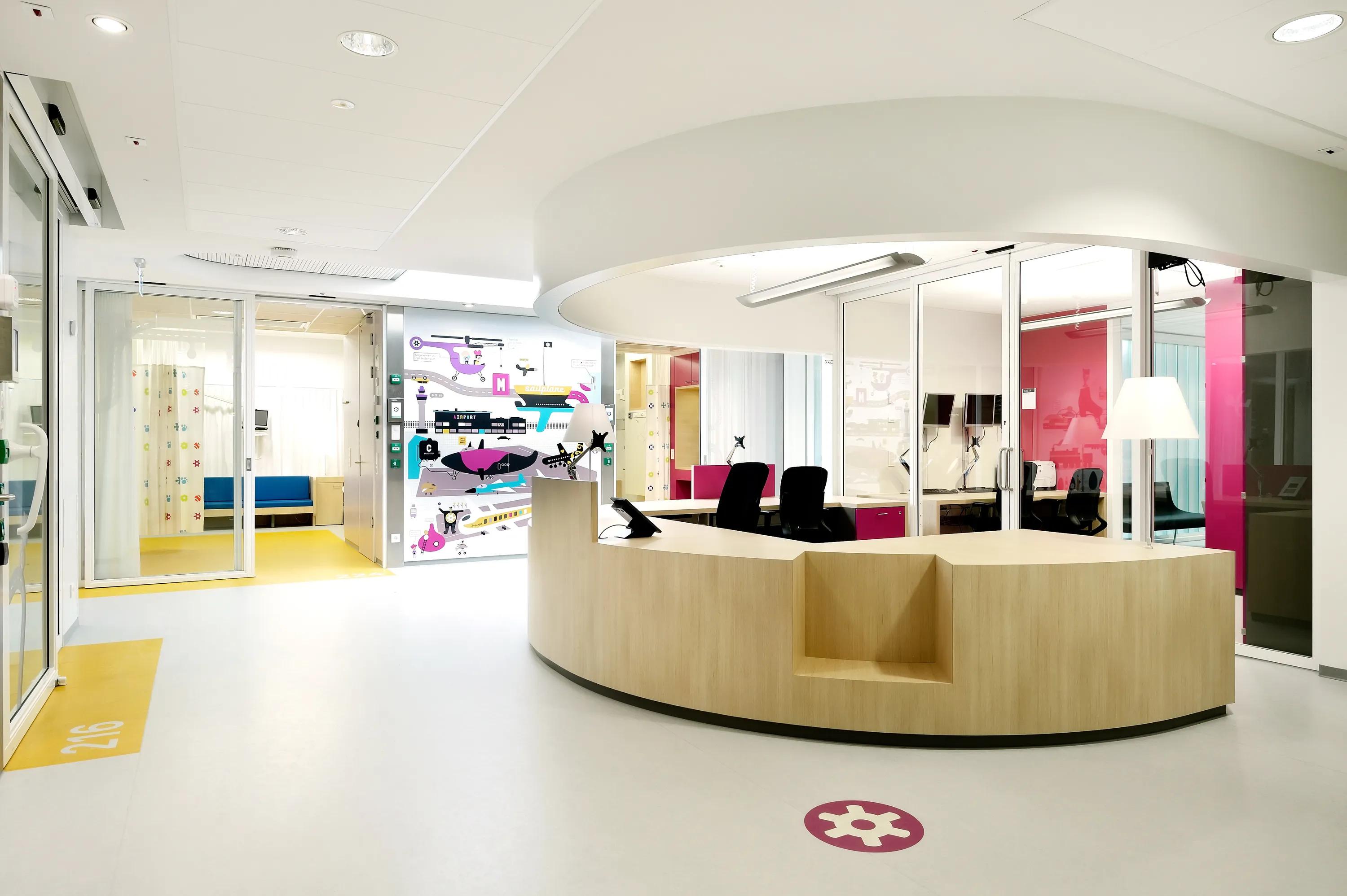 Children’s hospital reception area with a curved wooden desk, colourful wall graphics and a white acoustic ceiling with integrated lighting.