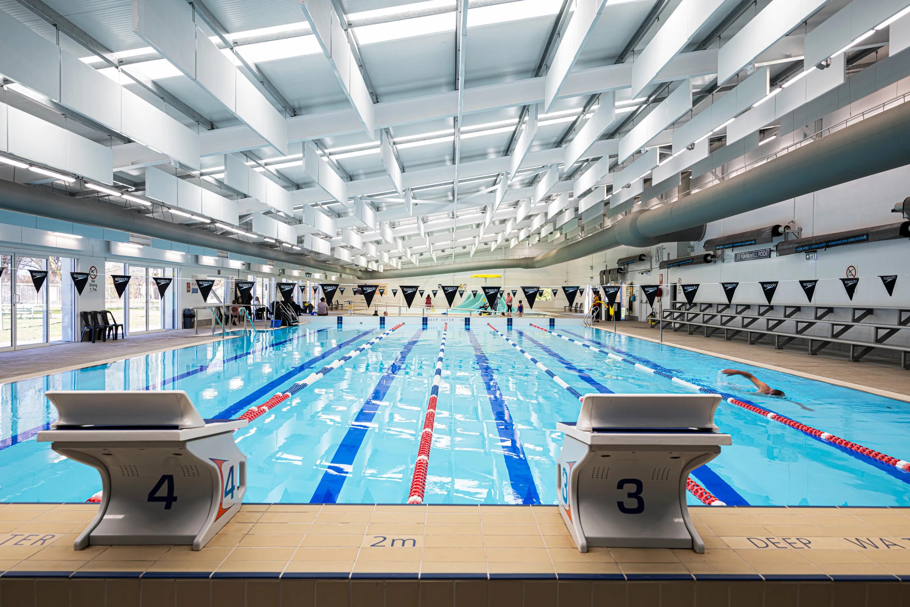 Indoor swimming pool with suspended white acoustic baffles across the ceiling, lane markings, starting blocks, and spectator seating along the poolside.