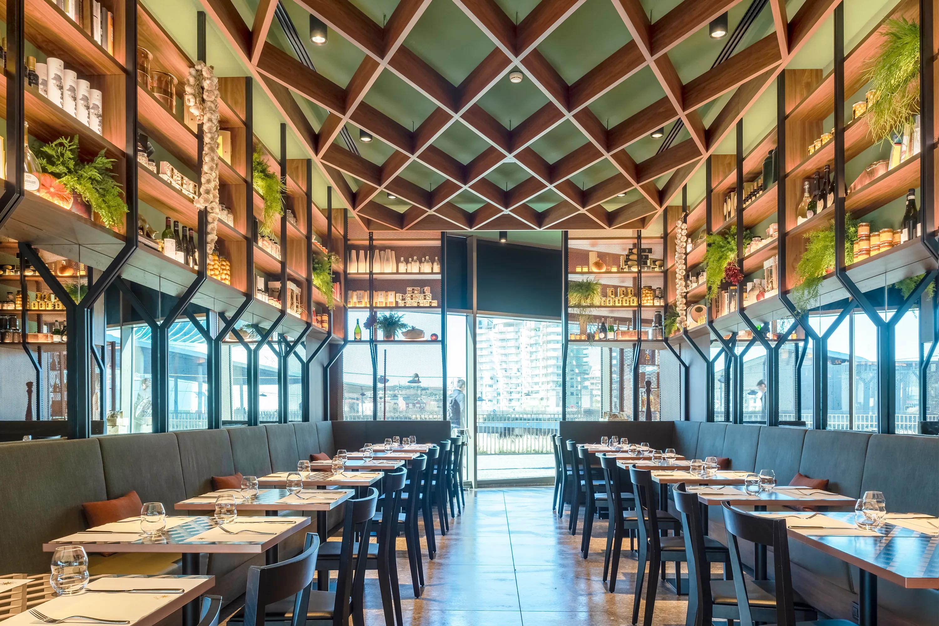 Modern restaurant interior with wooden shelves, green ceiling, and neatly arranged tables. Large window at the back offers a view outside.