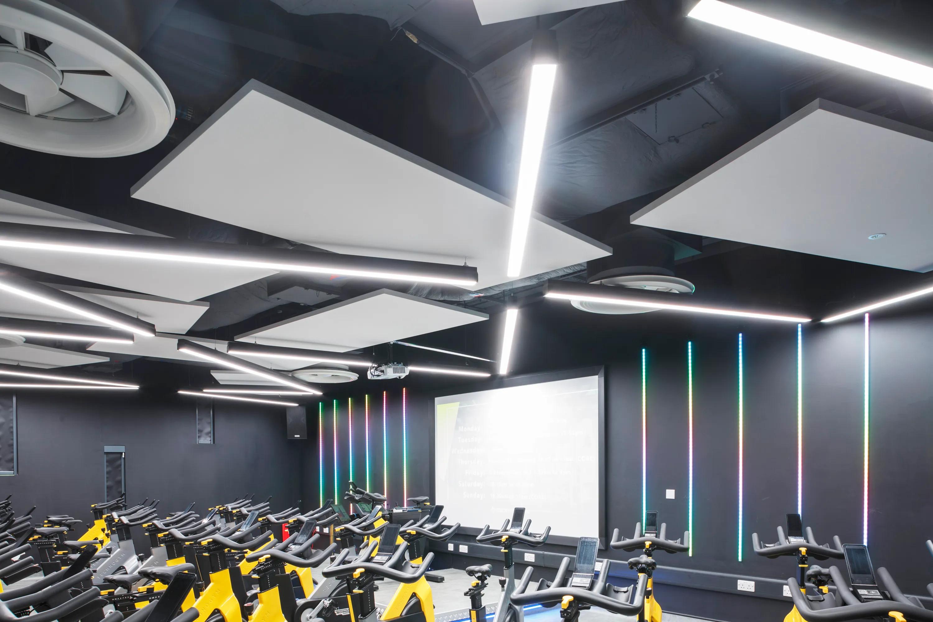 Modern indoor cycling studio with white floating ceiling panels, rows of stationary bikes, vibrant LED light strips, and geometric ceiling panels.