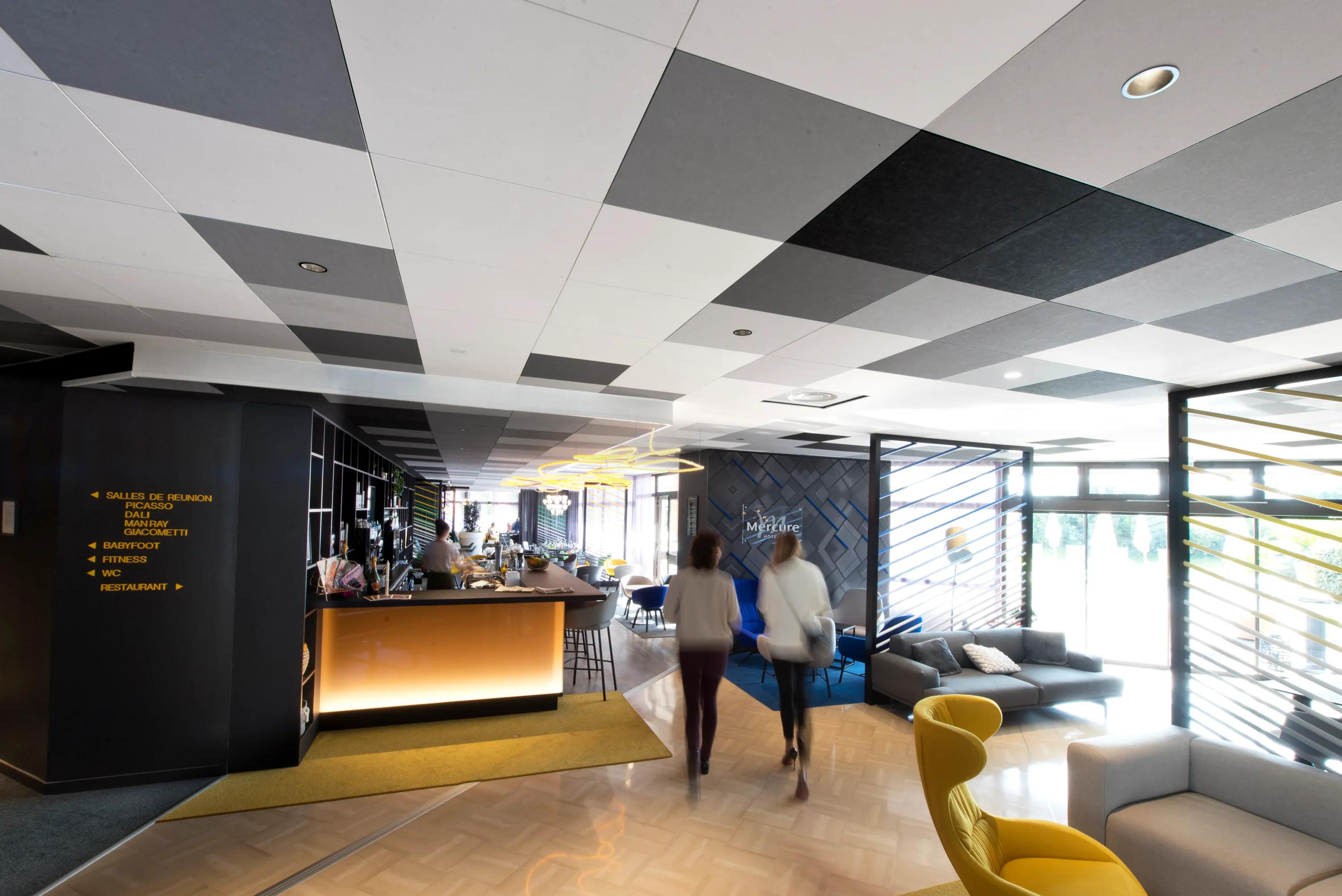Hotel bar and lounge with a black and white checkerboard acoustic ceiling, illuminated bar counter, seating areas, and large windows bringing daylight into the space.