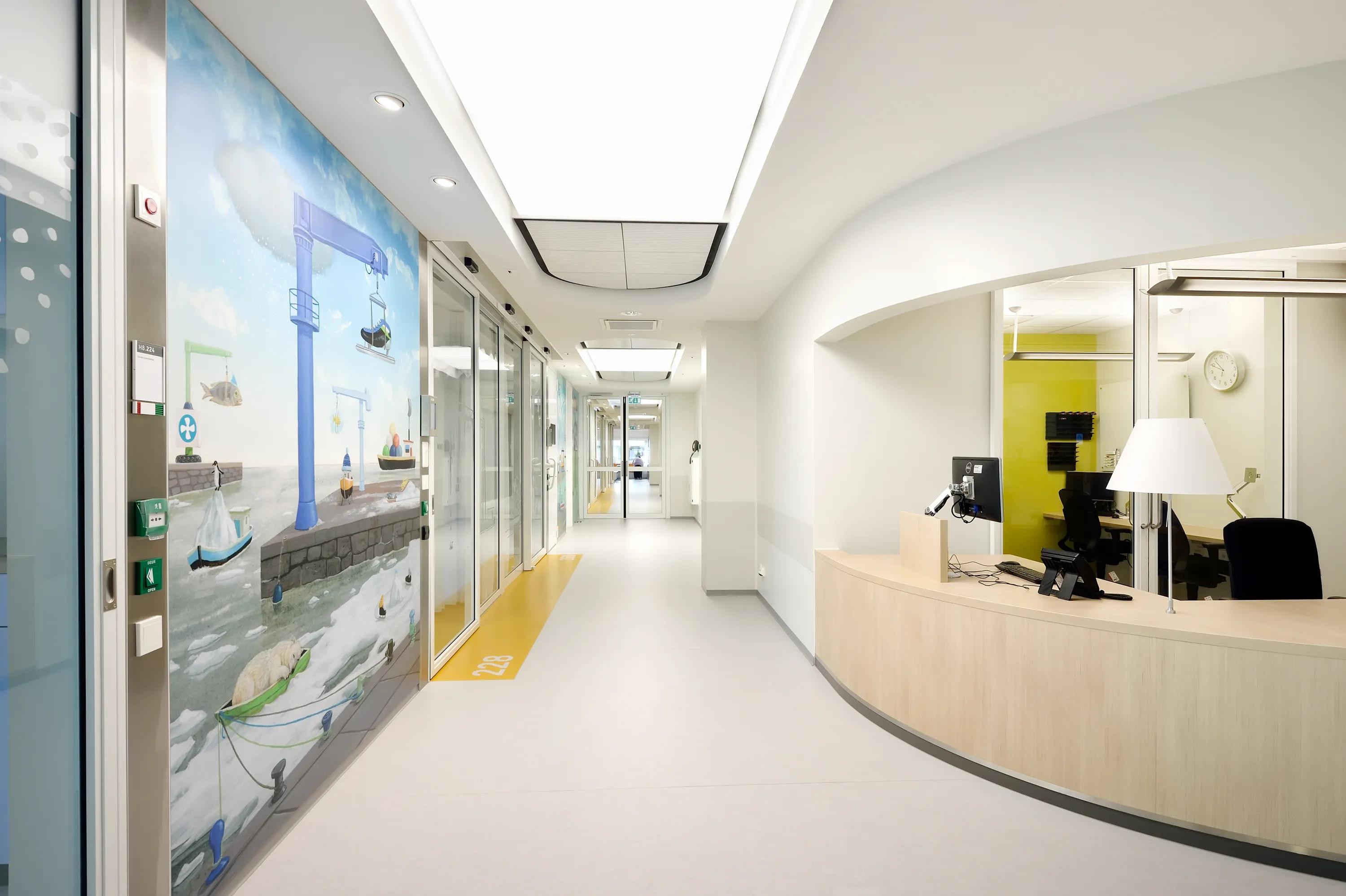 Hospital corridor featuring a nurse station with wall art, a curved reception desk and a large illuminated acoustic ceiling panel running down the hallway.
