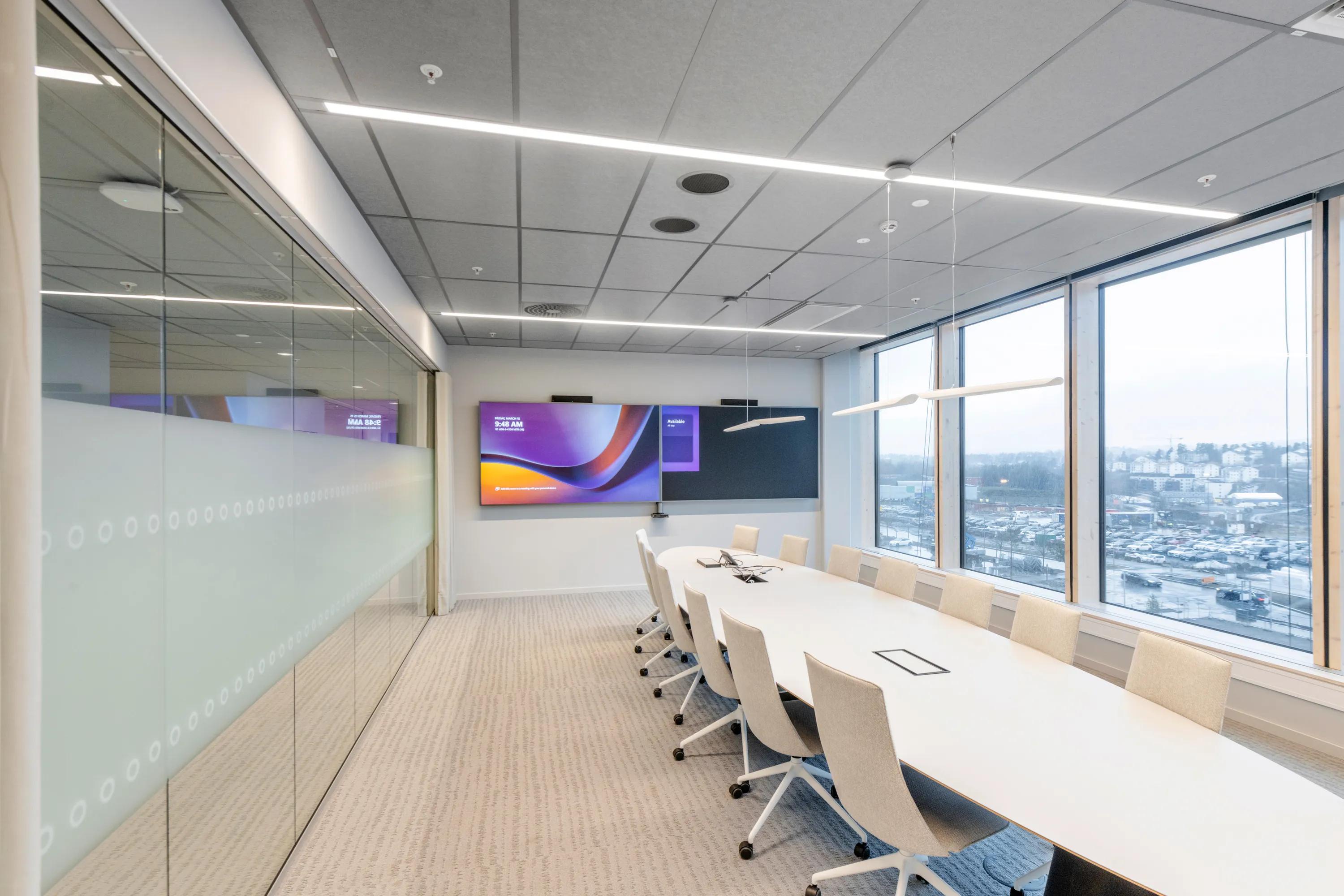Modern conference room with a long meeting table, large digital display, and a grey acoustic tile ceiling with integrated linear lighting above the space.
