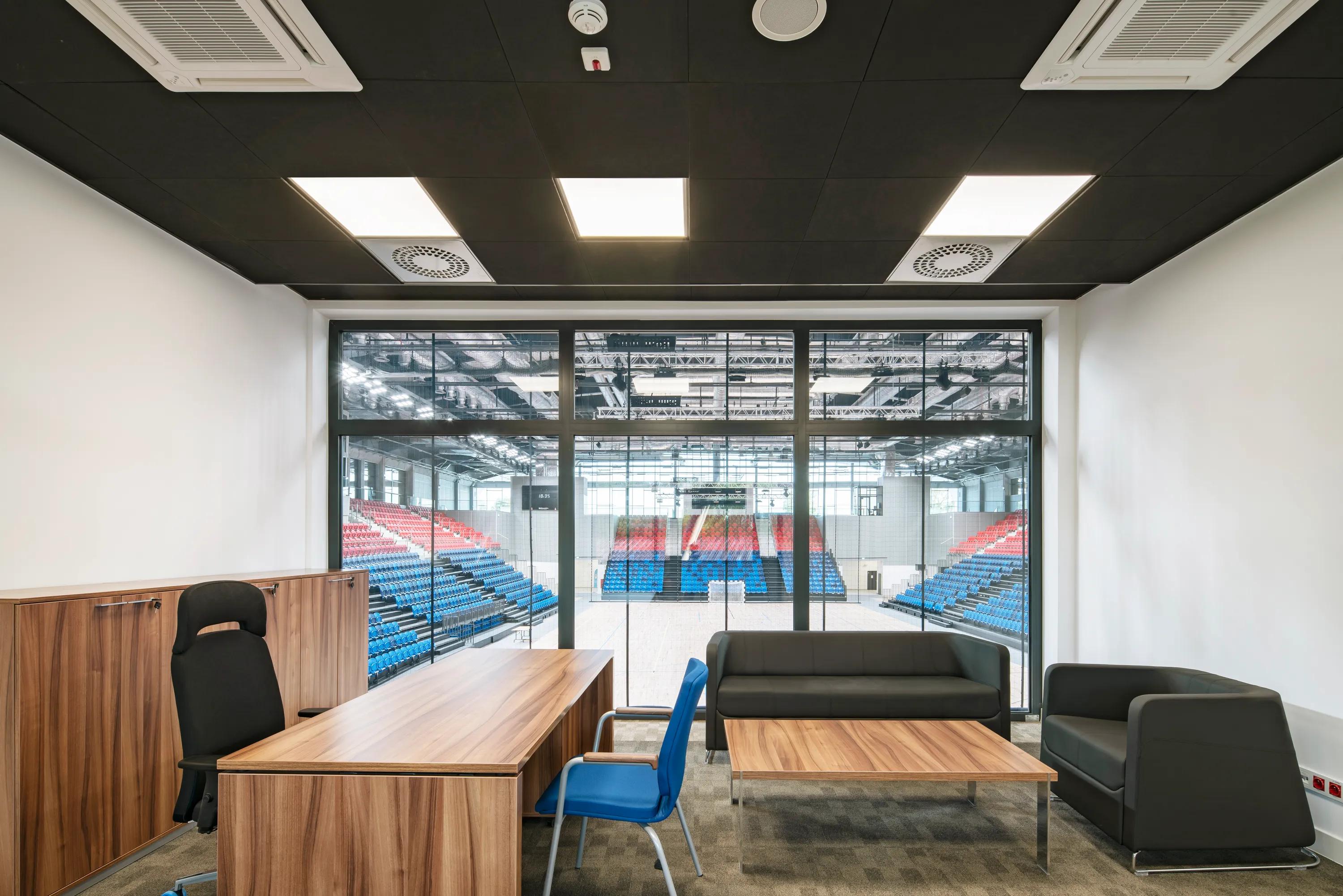Office with black suspended ceiling, wooden desk, chair, and black sofas overlooks an indoor sports arena with red and blue seating.