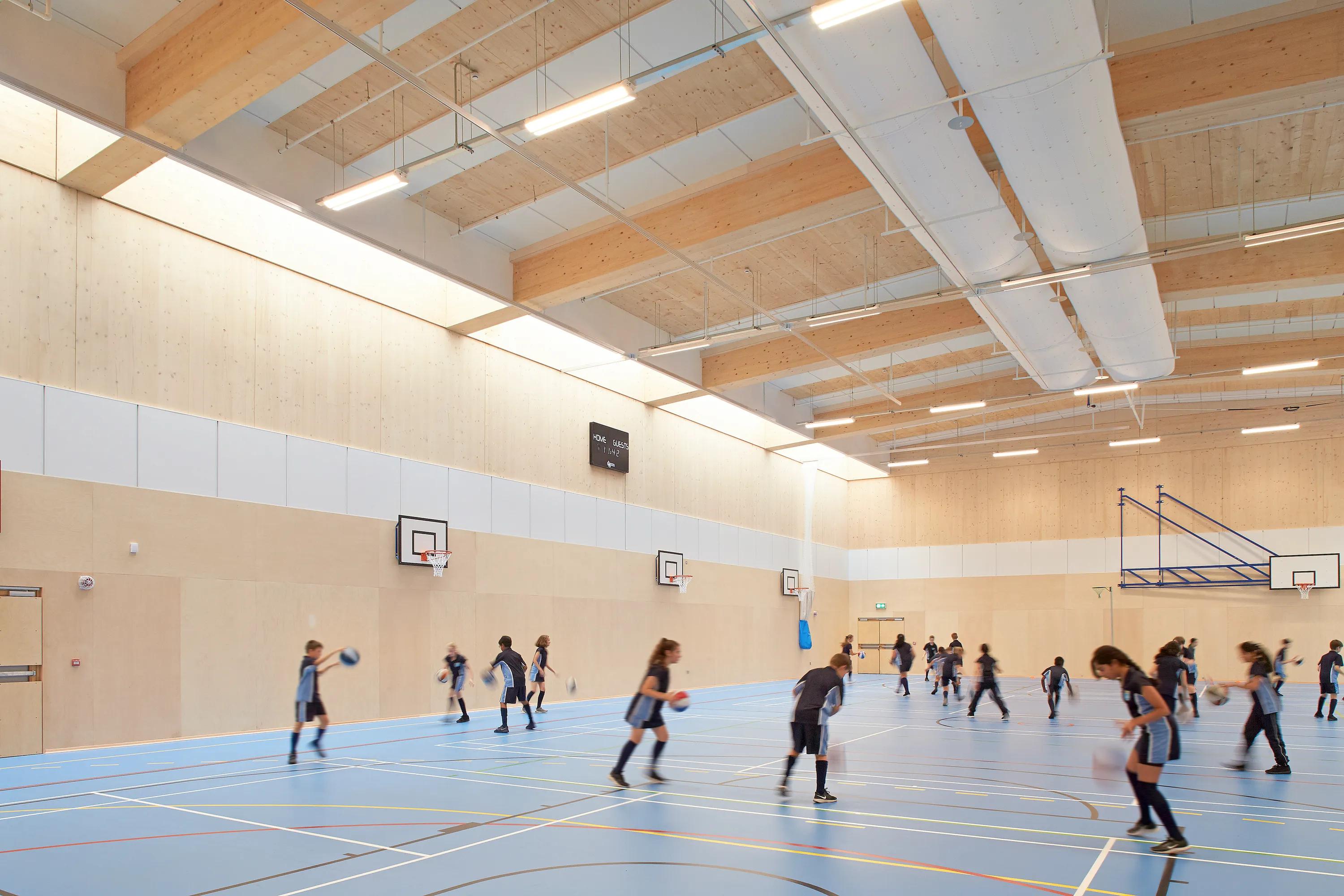 School sports hall with a timber structure, exposed beams and ventilation ducts, where students play basketball beneath suspended acoustic ceiling panels and linear lighting.