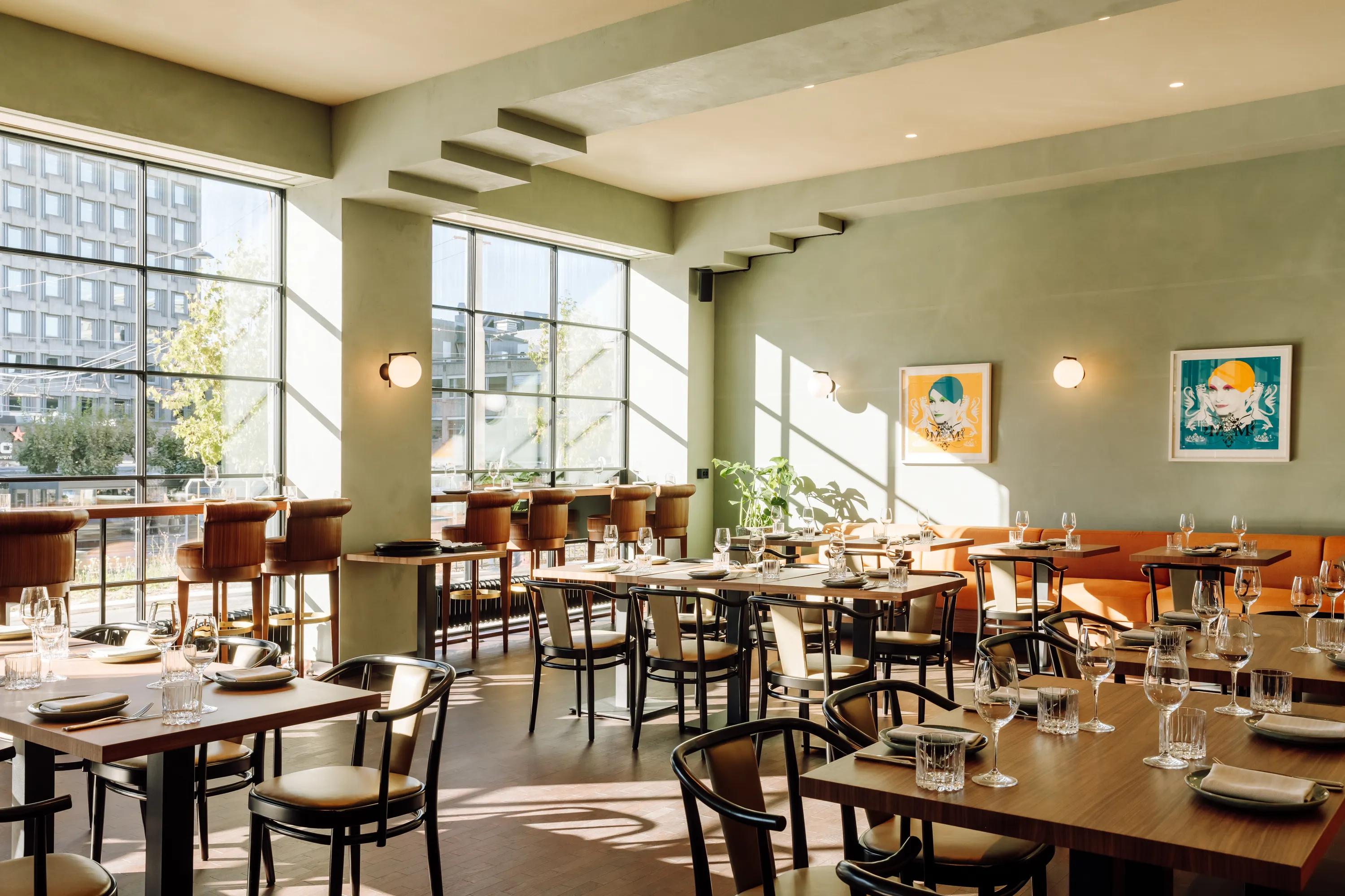 A bright, modern restaurant featuring a seamless monolithic acoustic ceiling in a soft neutral tone. The space includes large steel-framed windows, green plastered walls, warm wooden tables, bar seating, and framed artwork, with sunlight casting patterned shadows across the dining area.