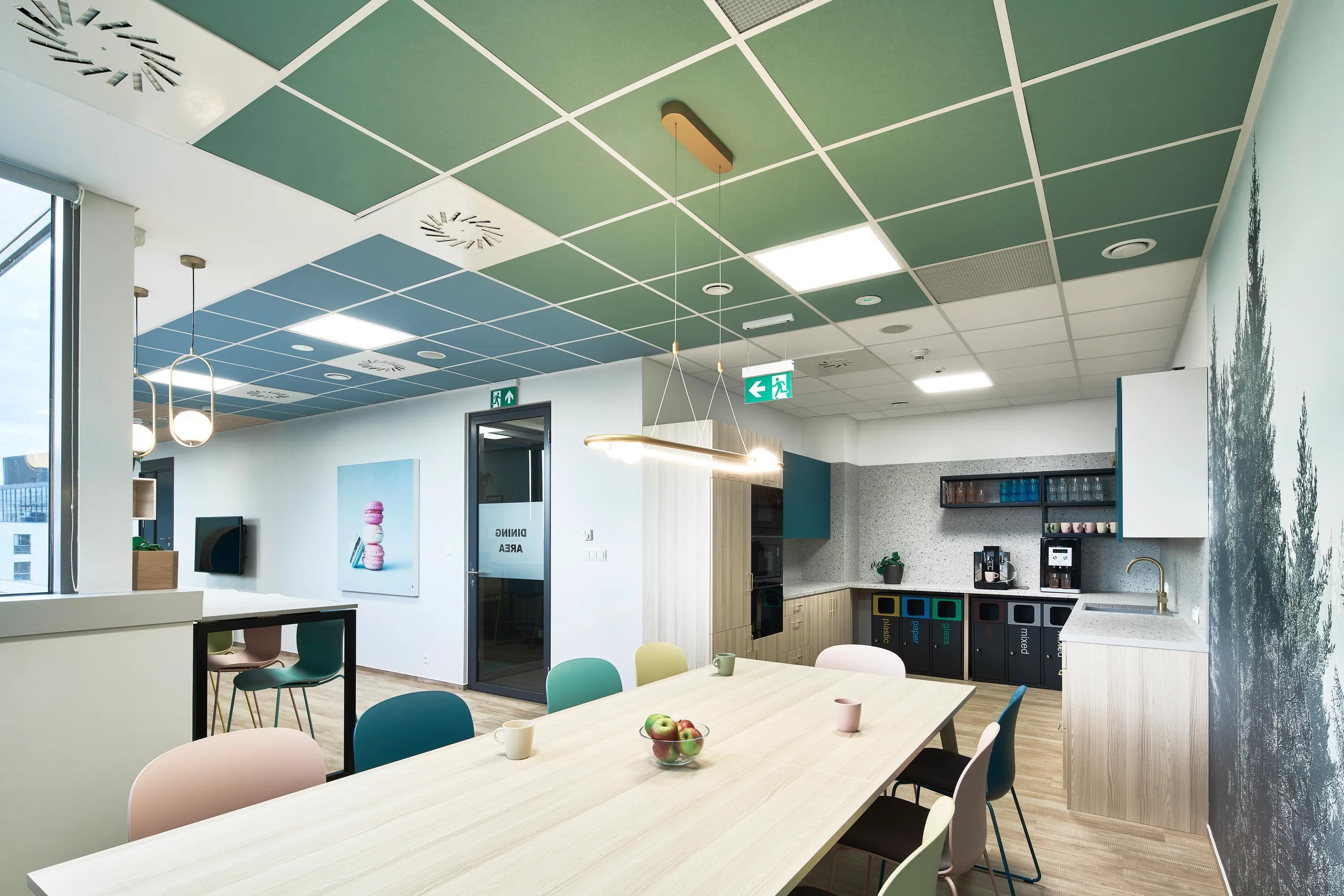 Modern office kitchen with a long wooden table, colorful chairs, and a kitchenette. Green and blue ceiling panels enhance the space.