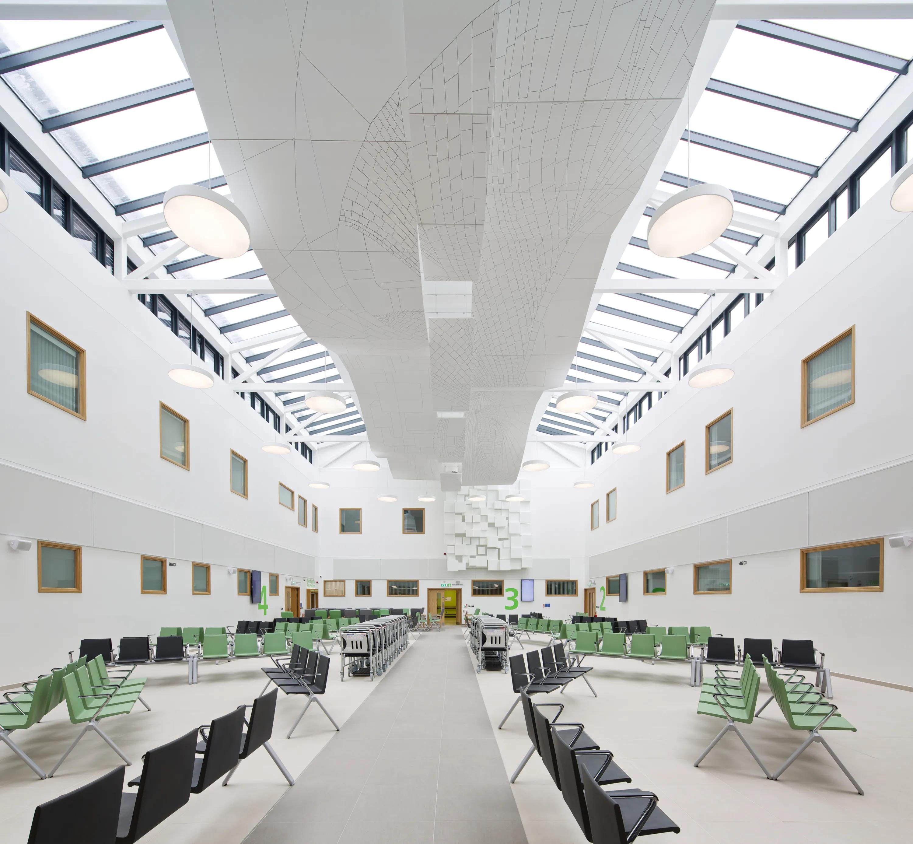 Spacious atrium with a floating raft ceiling and skylights, green and black seating areas, and geometric ceiling installations; modern and bright ambiance.