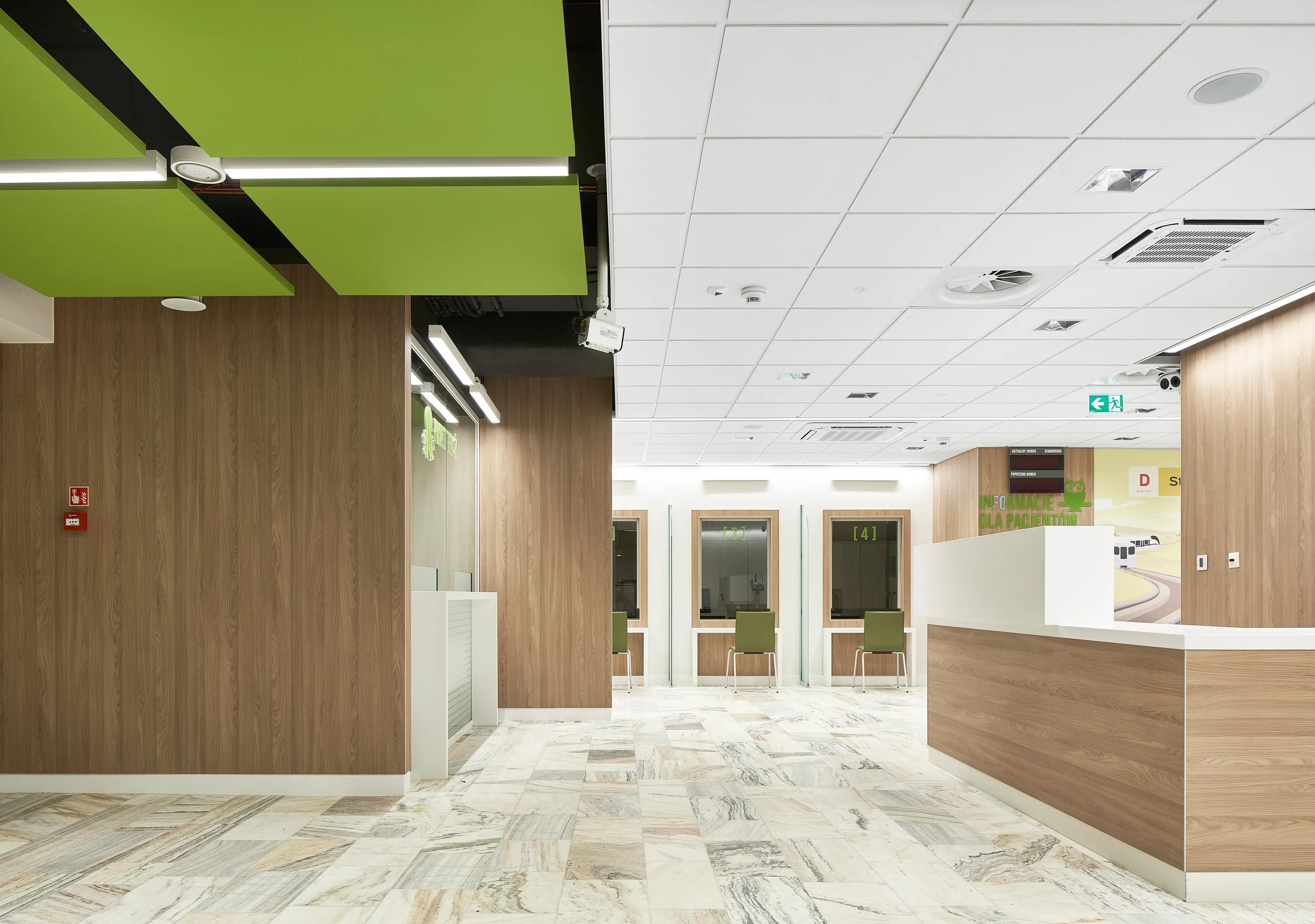Hospital reception area featuring a white acoustic ceiling with integrated services and suspended green acoustic islands, complemented by a wood-panelled desk and green waiting benches.