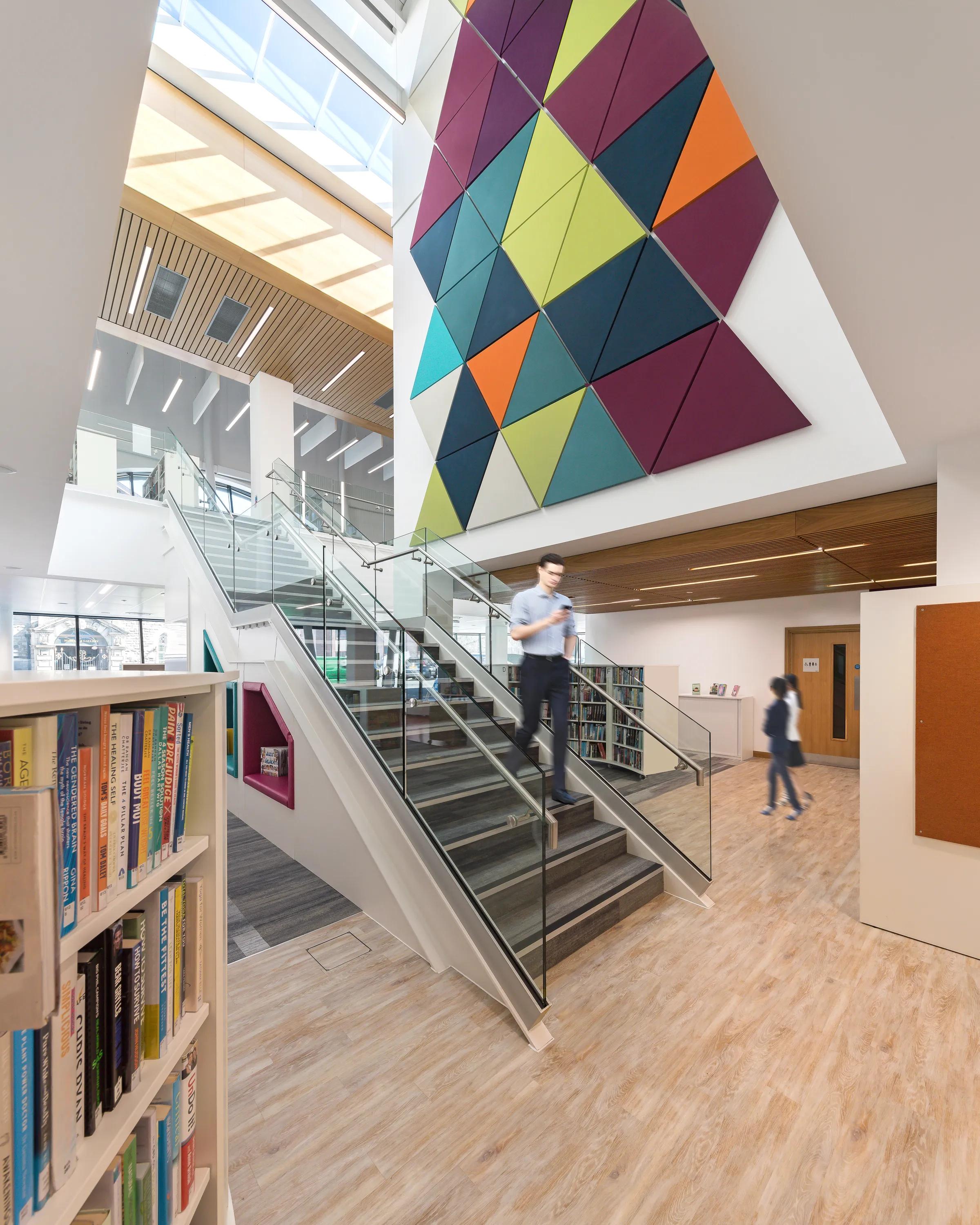Library atrium with a staircase, bookshelves, and a large colourful geometric acoustic wall panel installation beneath a skylight.
