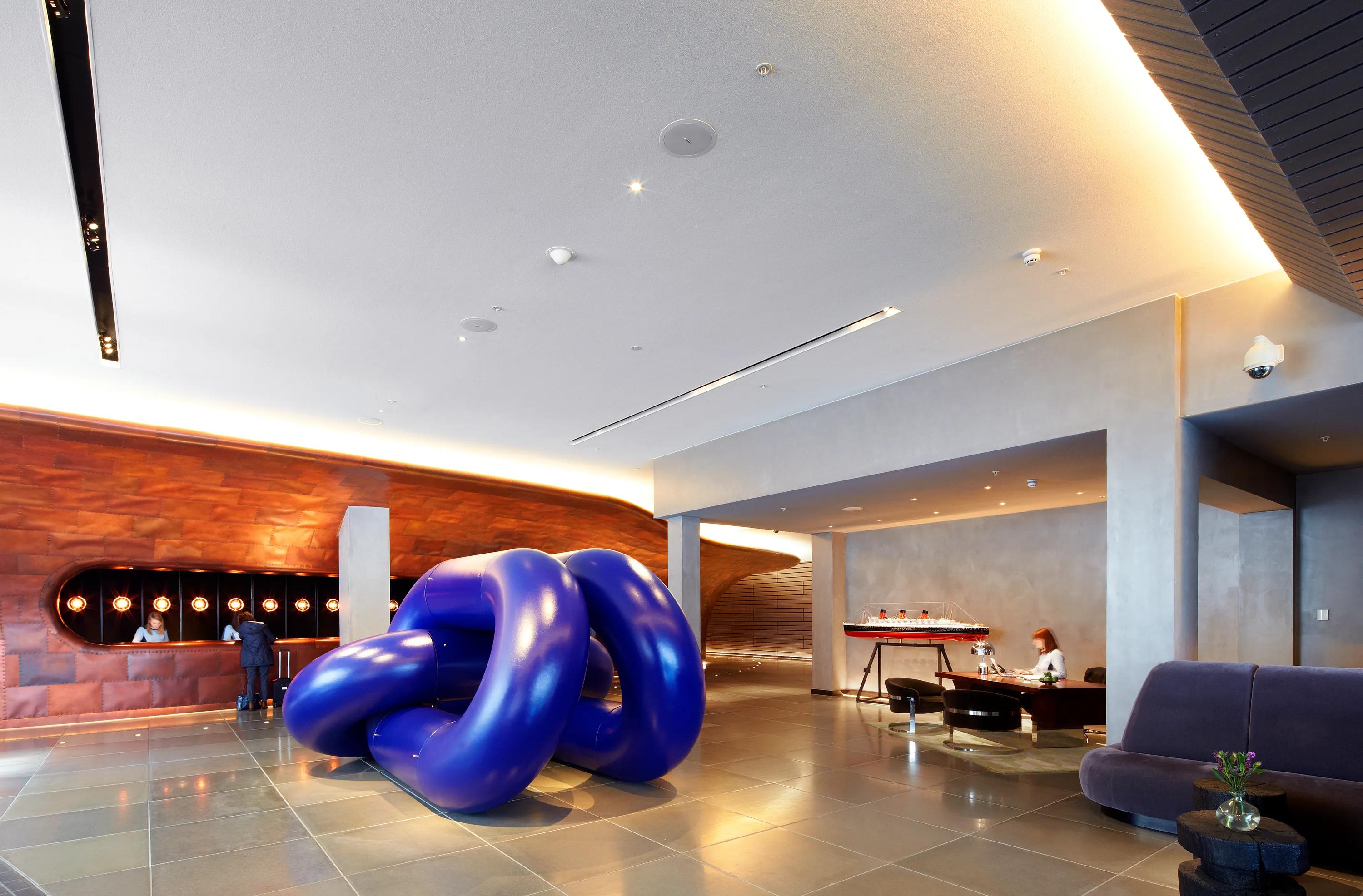 Hotel reception lobby with a smooth monolithic acoustic ceiling, curved copper-clad reception desk, and a large blue sculptural installation positioned in the centre of the space.