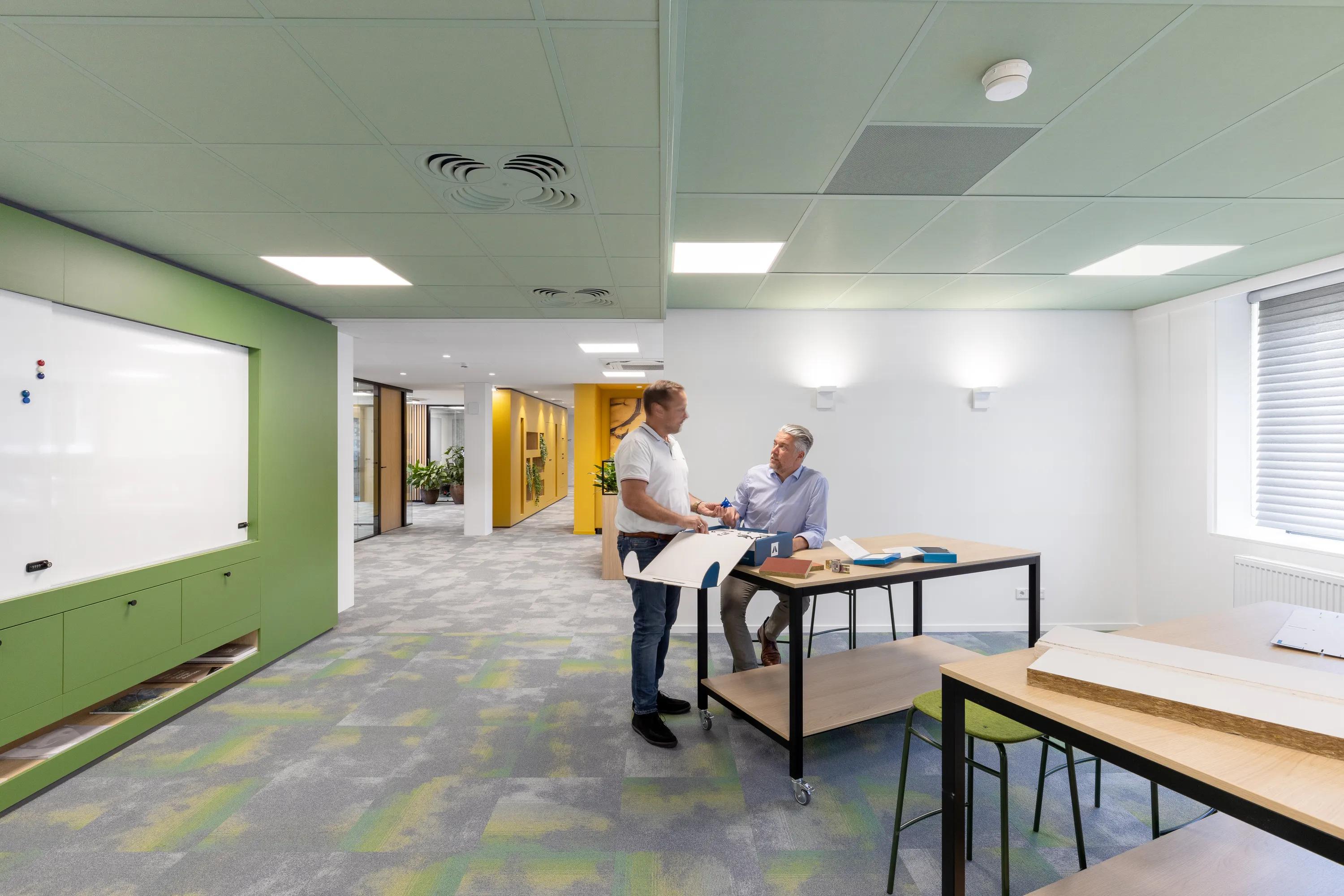 Two people collaborating at a standing desk in a modern office with green accents, whiteboard, and glass partitions.