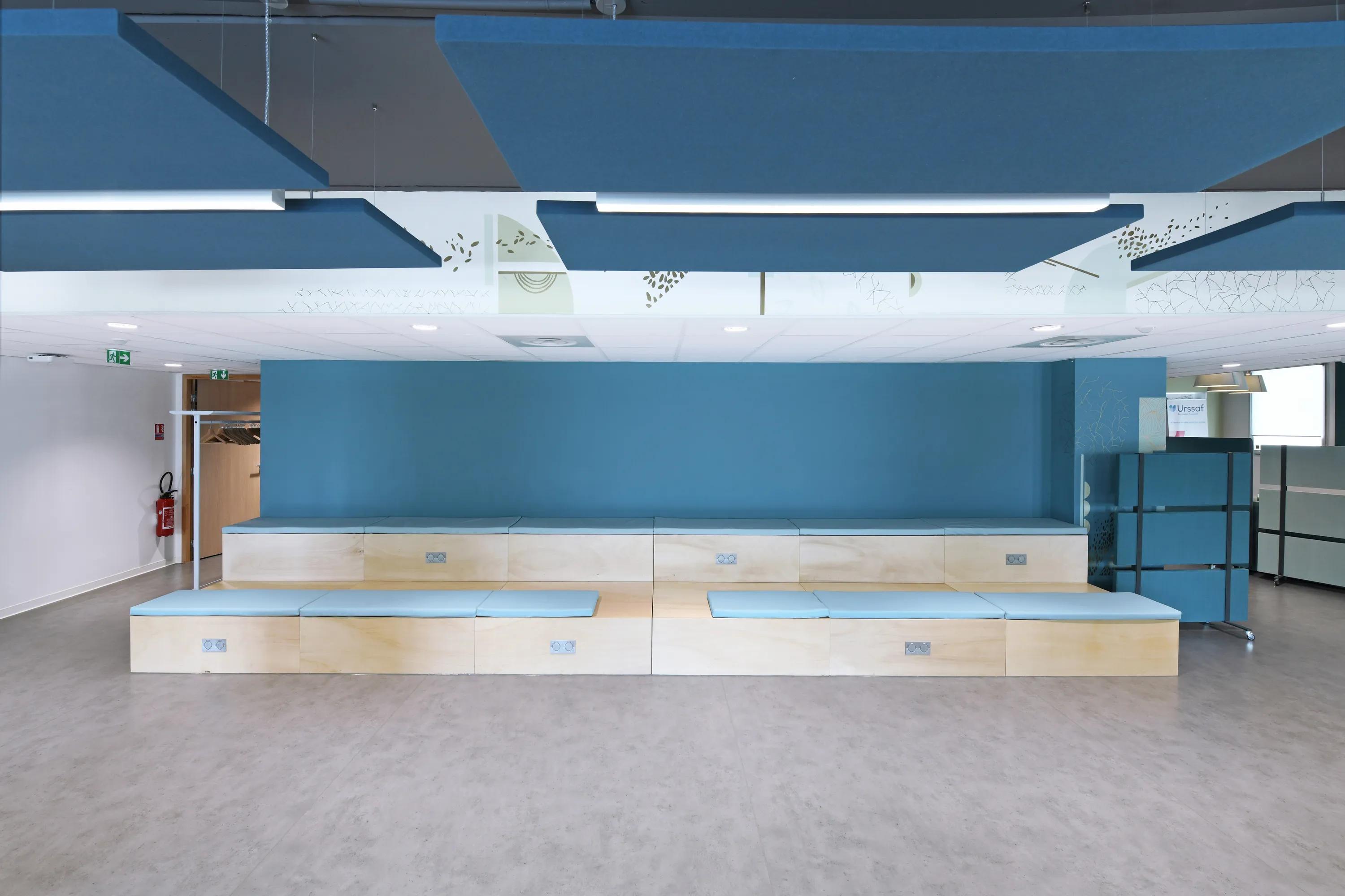 Modern waiting area with blue floating ceiling panels, light wood benches, blue accent wall, and decorative ceiling panels. Door and emergency exit signs are visible.