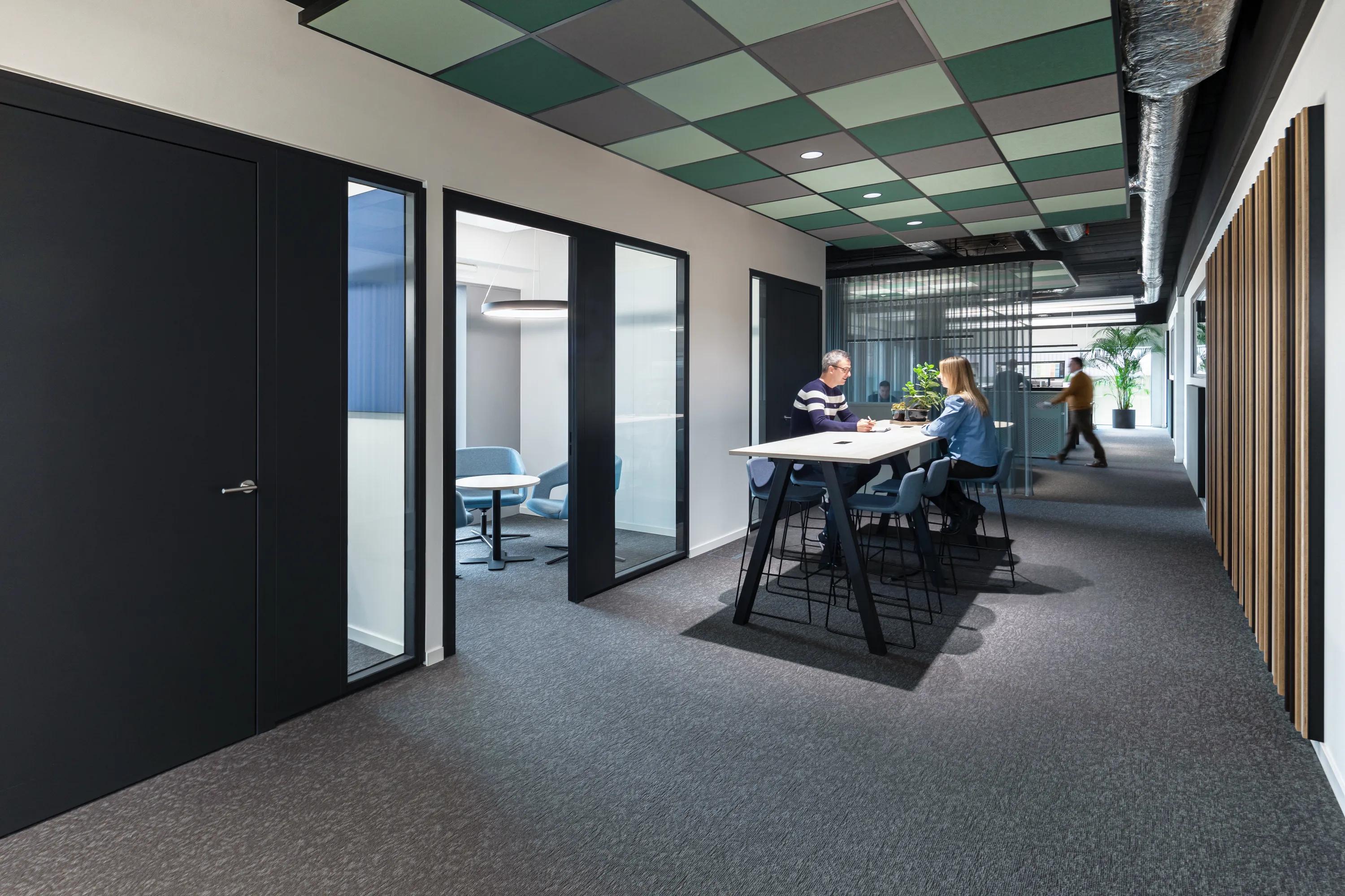 Modern office hallway with black doors, glass-walled meeting rooms, and a high table. Two people are seated, engaged in conversation.