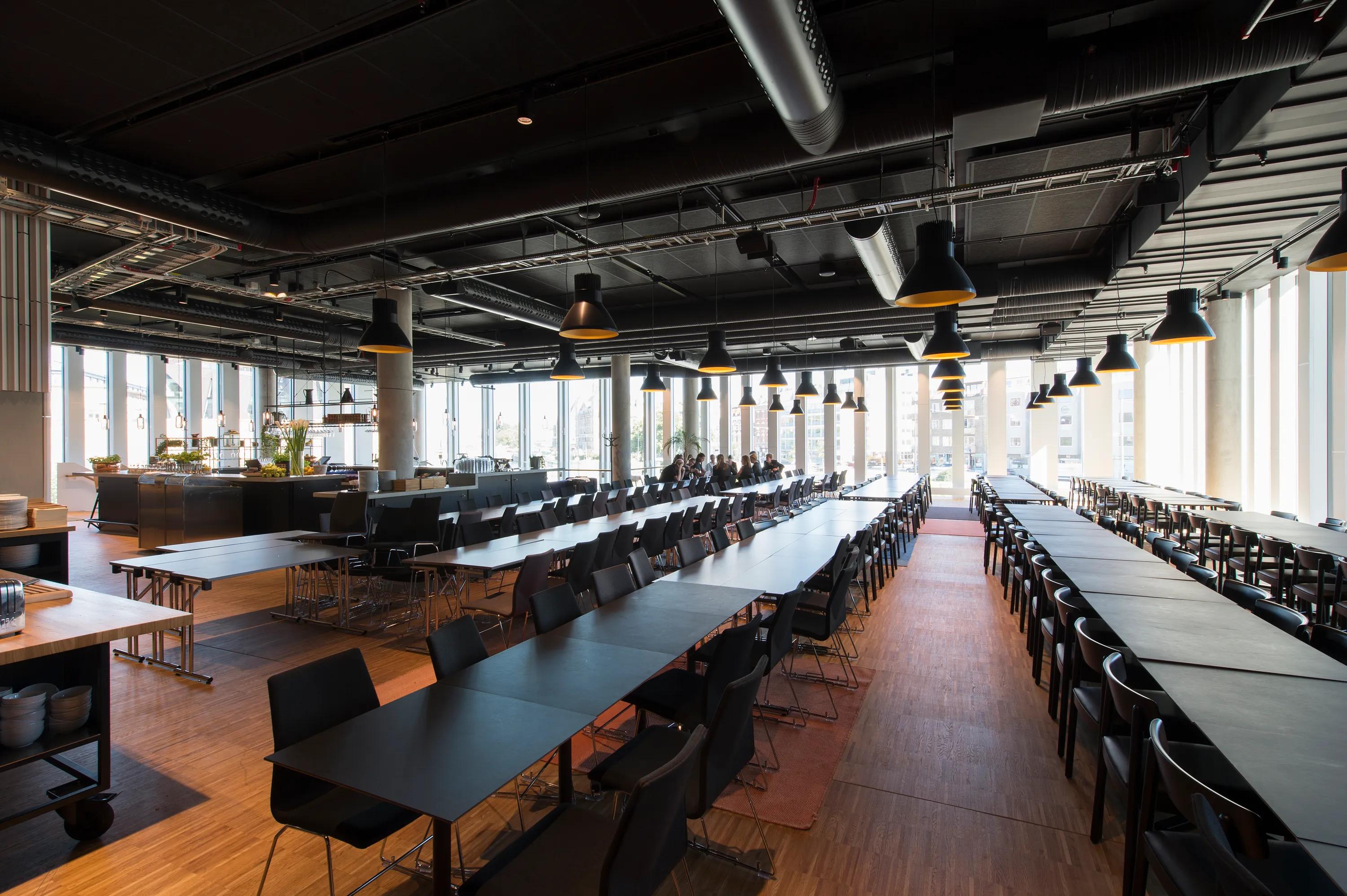 Spacious, modern cafeteria with a black suspended ceiling, black tables and chairs, large windows, and overhead lighting, featuring a clean, industrial design.