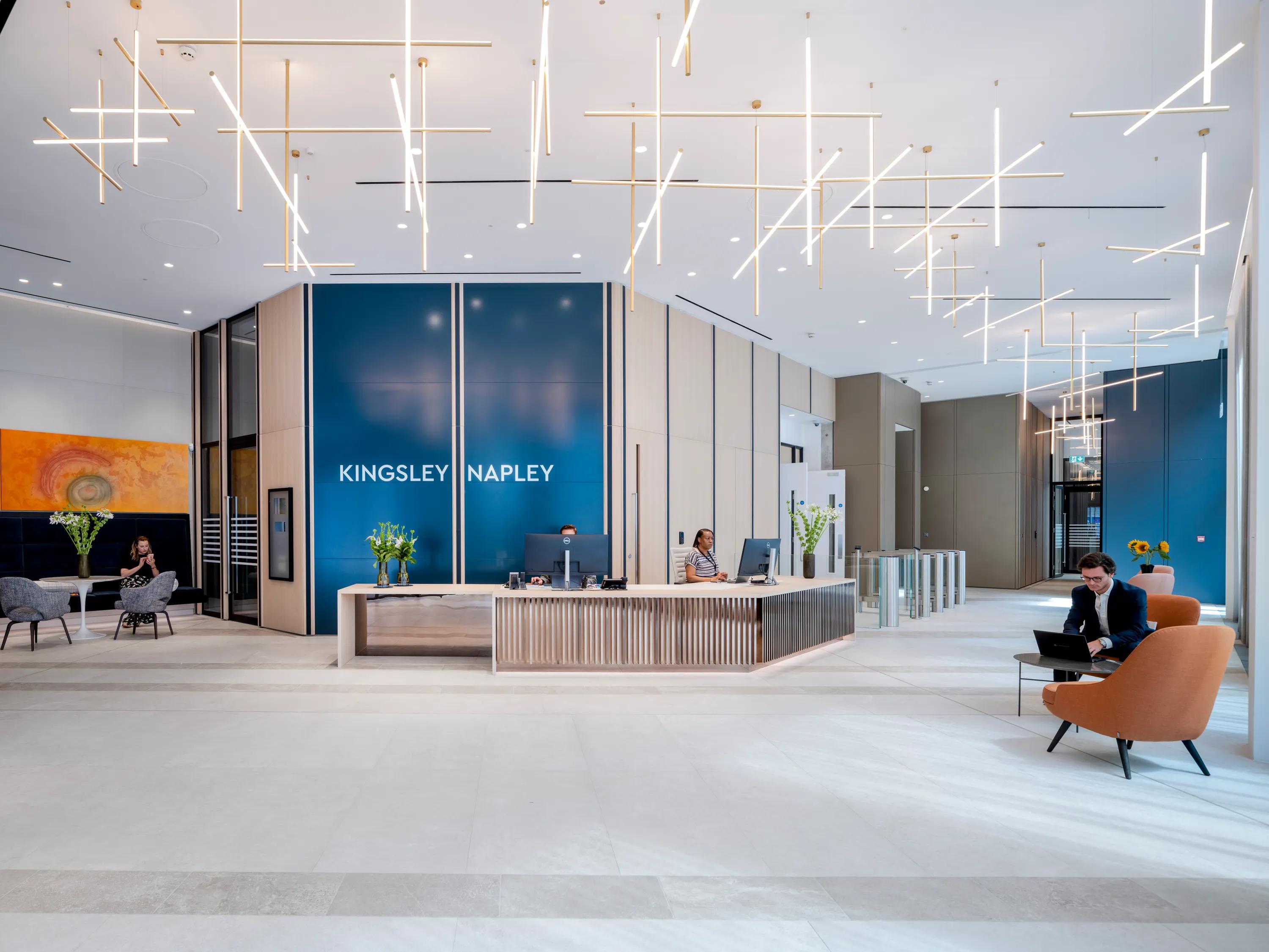 Modern office reception with a seamless white office ceiling, sleek desk, blue panels, seated visitors, and glowing linear ceiling lights.