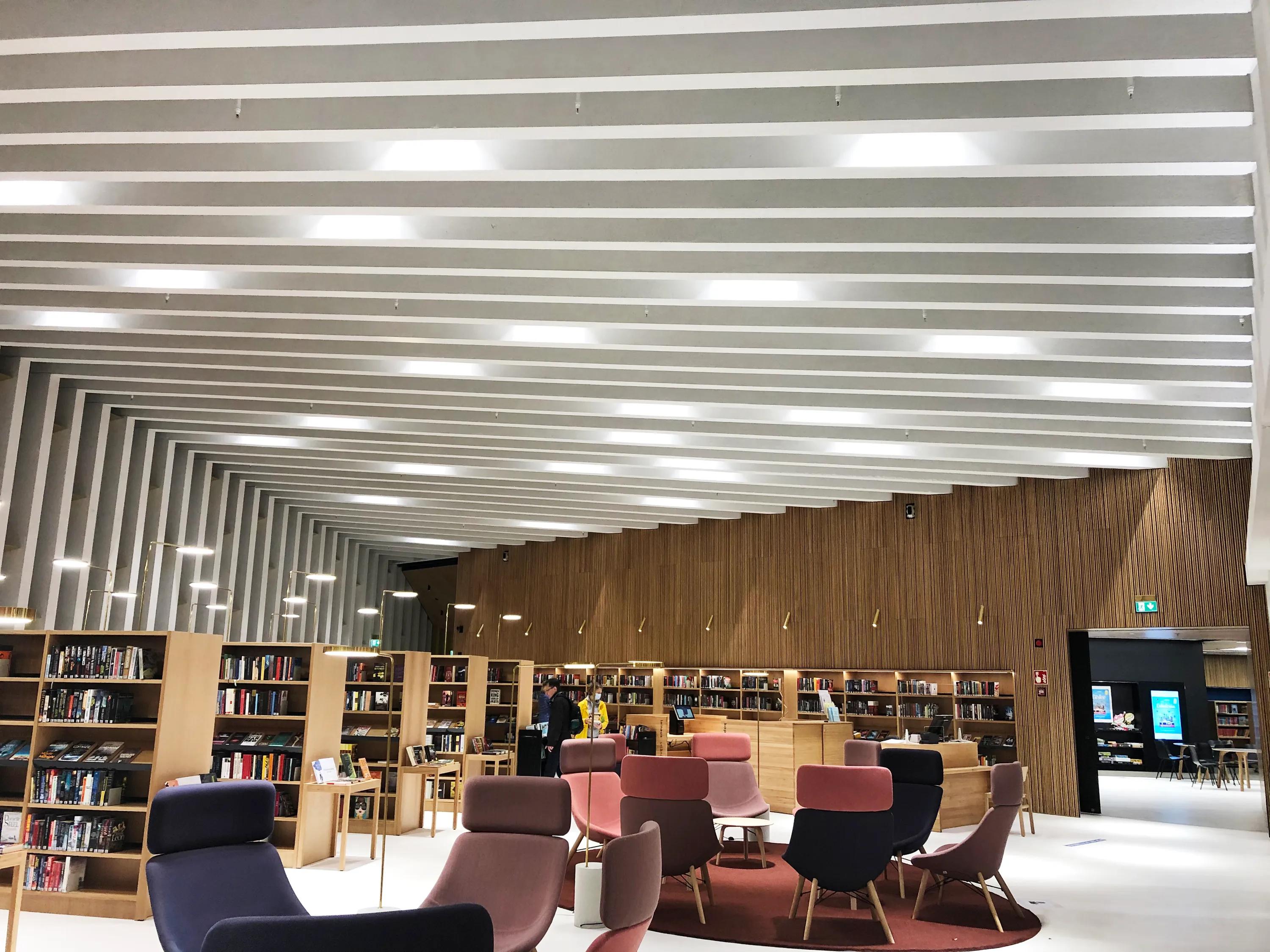 A spacious library interior featuring rows of long white acoustic baffles across the ceiling, complemented by timber bookshelves and colourful lounge chairs