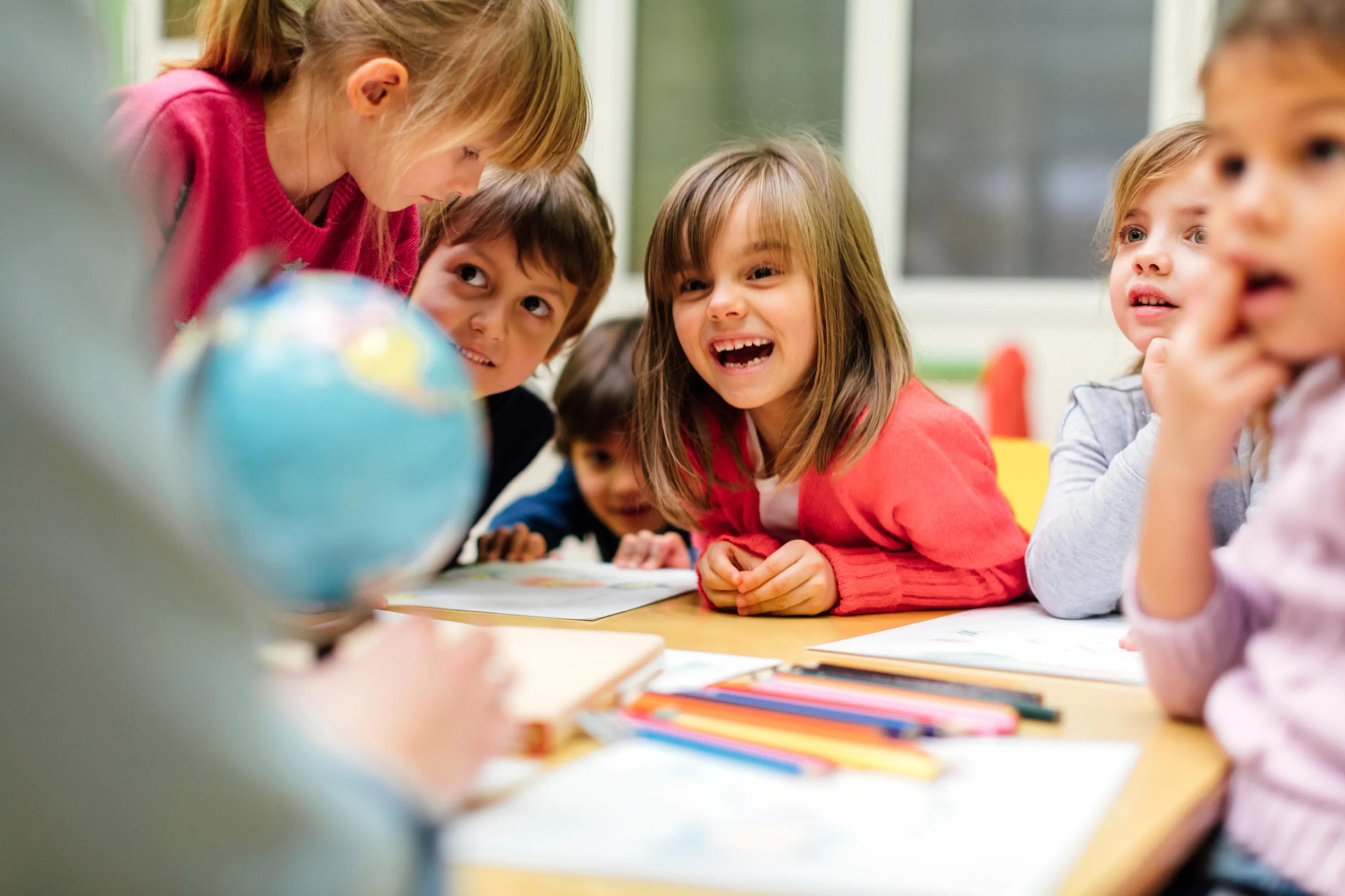 A group of young children sit around a classroom table, smiling and leaning in with curiosity as they look at a small globe and work on drawings.