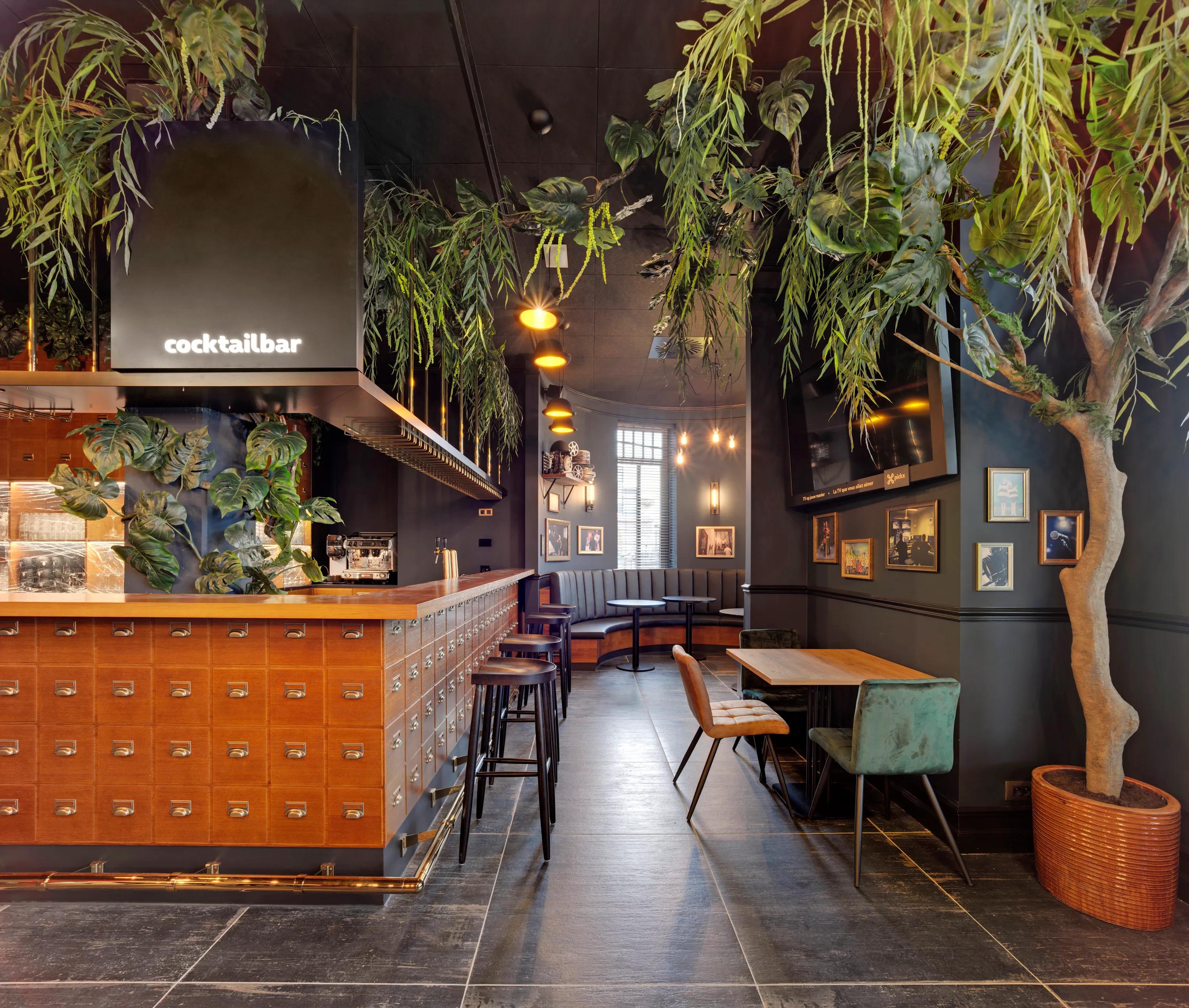 Stylish cocktail bar with black suspended ceiling, wooden counter, high stools, and lush greenery. Cozy seating area with upholstered chairs and framed art on walls.