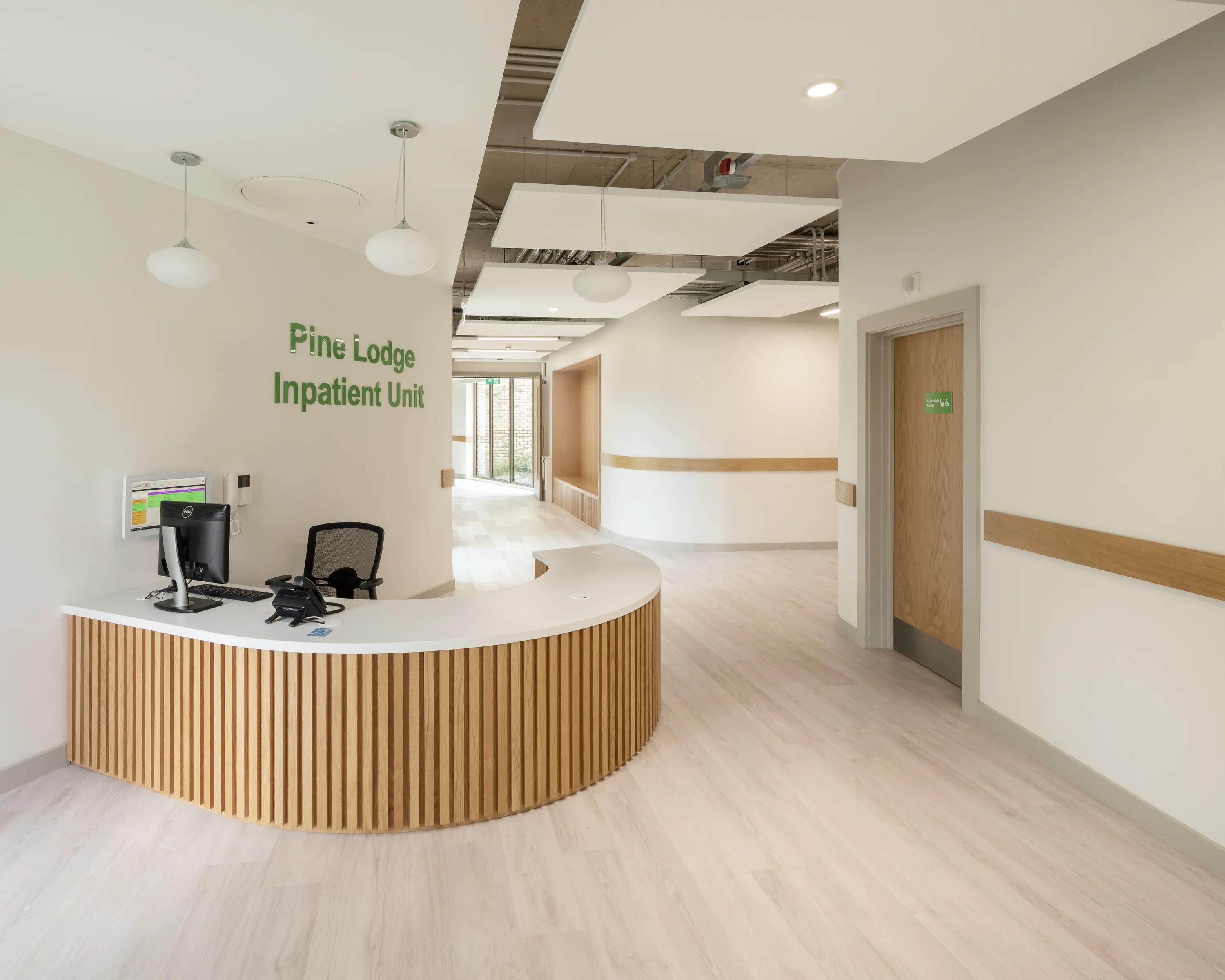 A welcoming inpatient unit reception with a curved wooden desk, pendant lighting and suspended white acoustic ceiling islands.