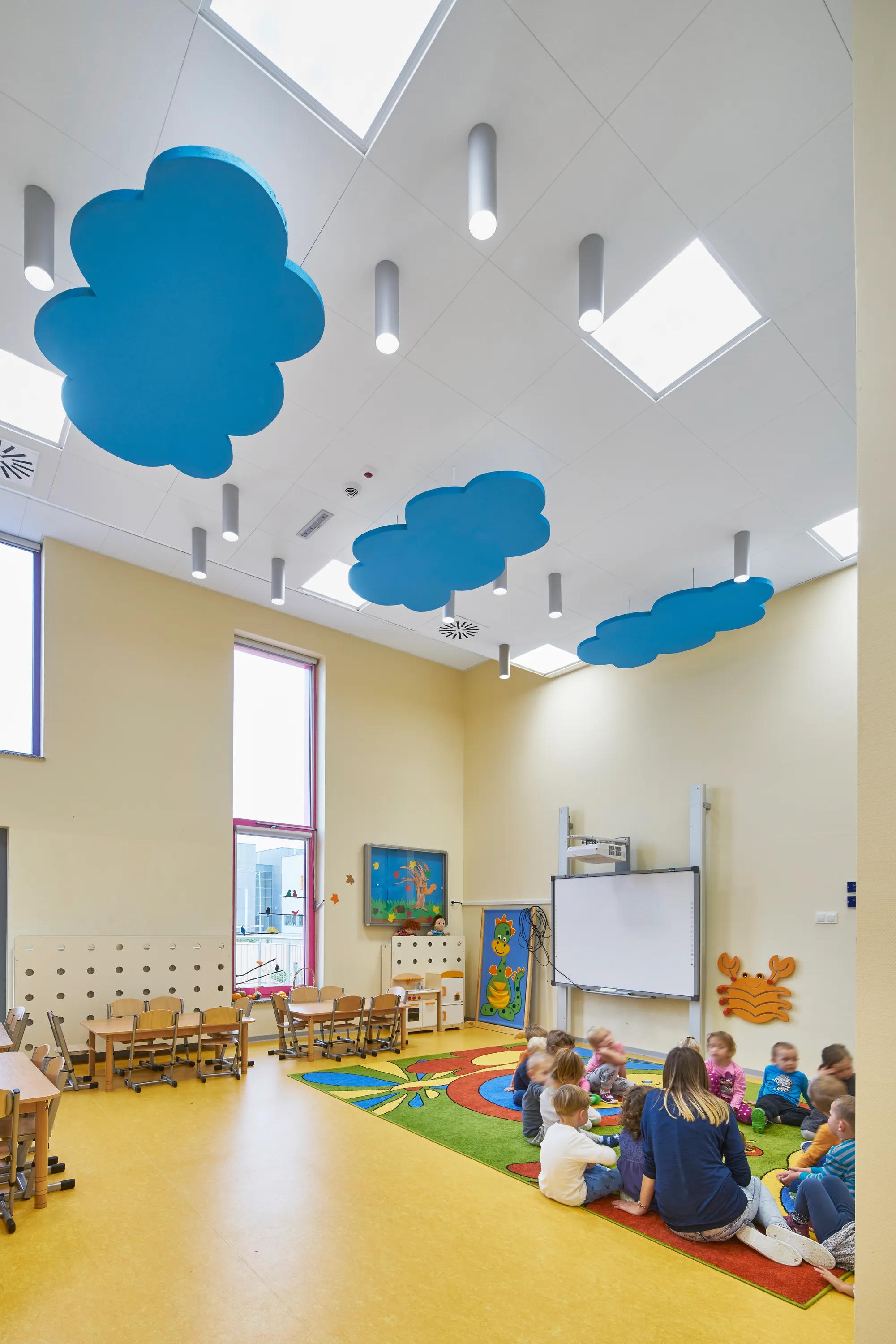 A playful nursery classroom featuring blue cloud-shaped acoustic ceiling islands suspended under white acoustic ceiling tiles and cylindrical pendant lights, with young children gathered on a colourful rug beneath high windows.